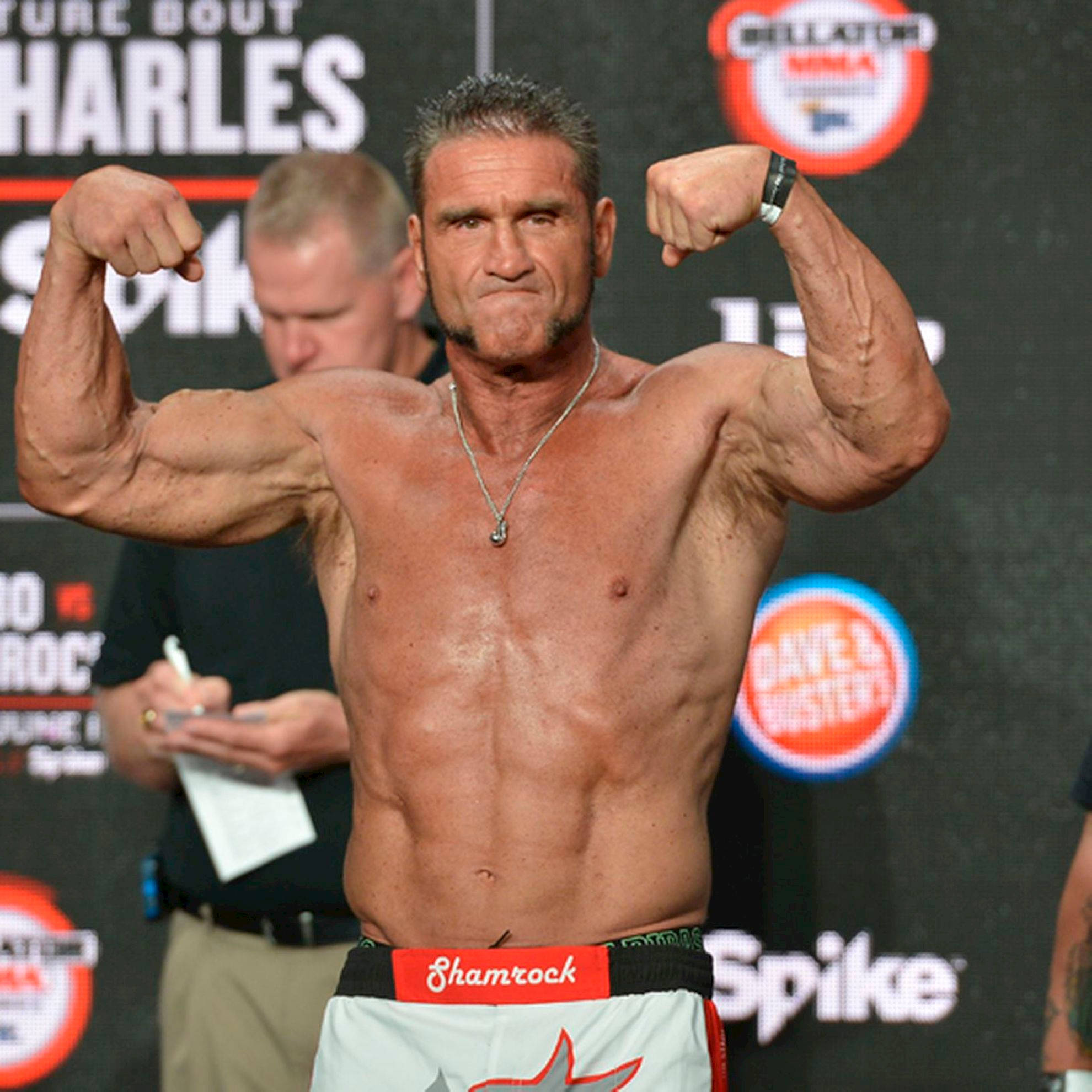MMA 桌布 壁紙 — 一位肌肉發達的拳手在稱重活動中彎曲手臂，身後有贊助商標誌。