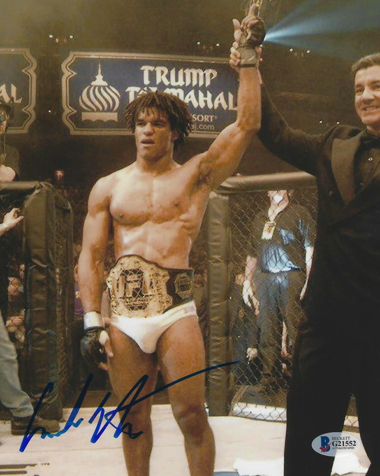 UFC 桌布 壁紙 — 一位肌肉發達的拳手穿著白色短褲和冠軍腰帶，在明亮的場館燈光下，站在MMA籠中勝利，一名西裝男子舉起他的手臂。