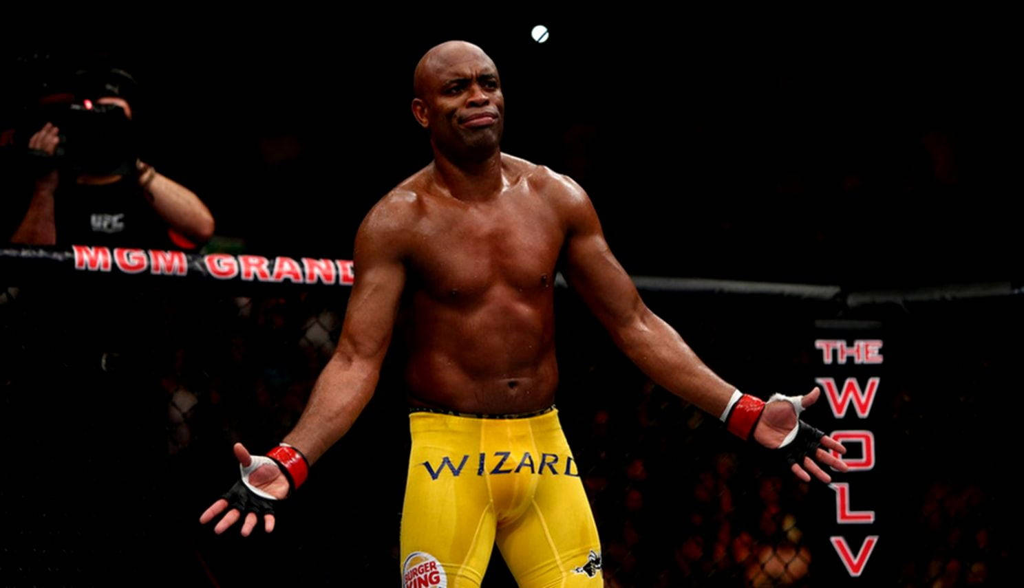 UFC 桌布 壁紙 — 一位肌肉發達的拳手穿著黃色短褲，在昏暗的八角籠中比賽後張開雙臂站立。