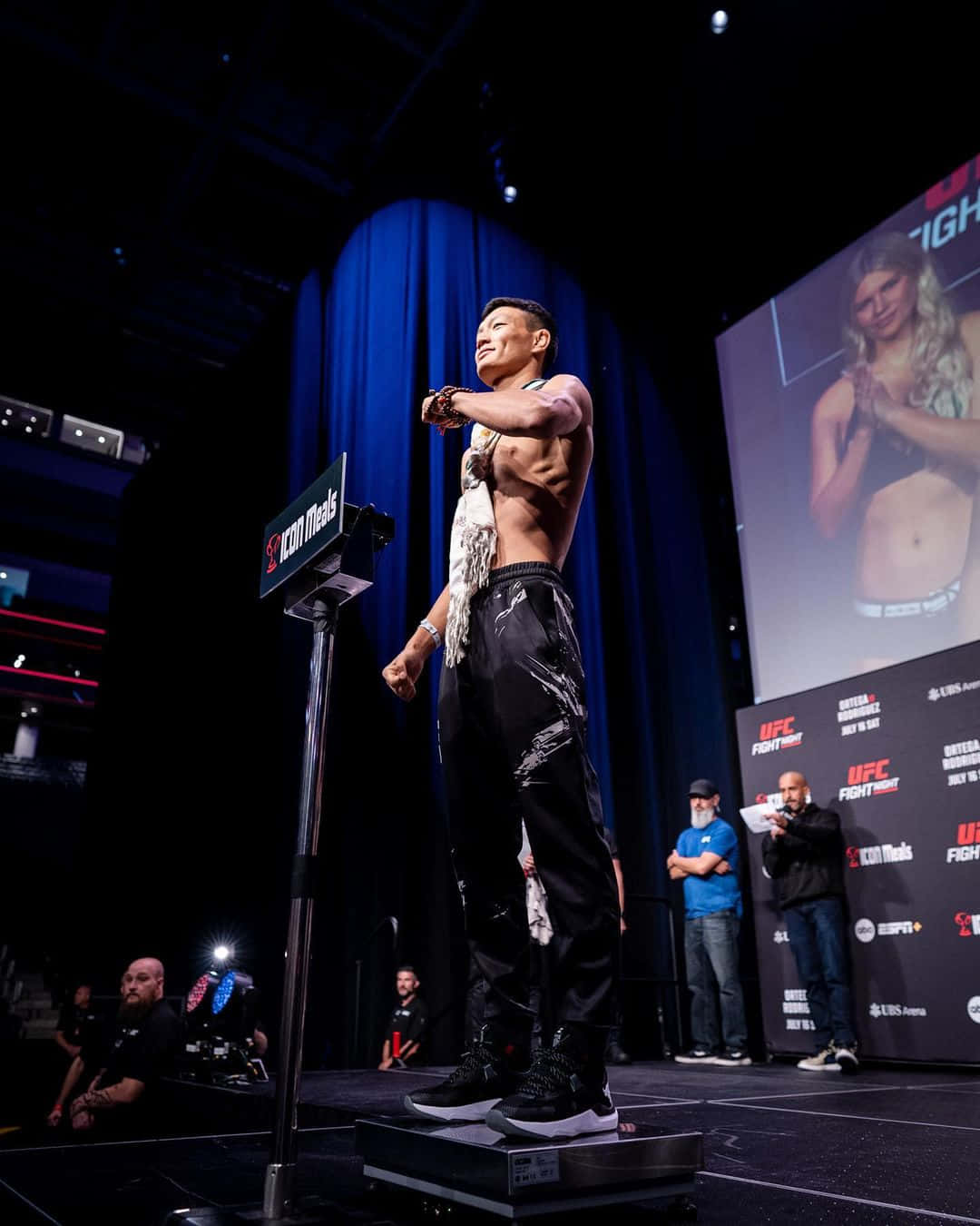 MMA 桌布 壁紙 — 一位肌肉發達的拳手自信地站在體重計上，在UFC稱重儀式中，明亮的舞台燈光下，身後的大螢幕顯示著他的對手。