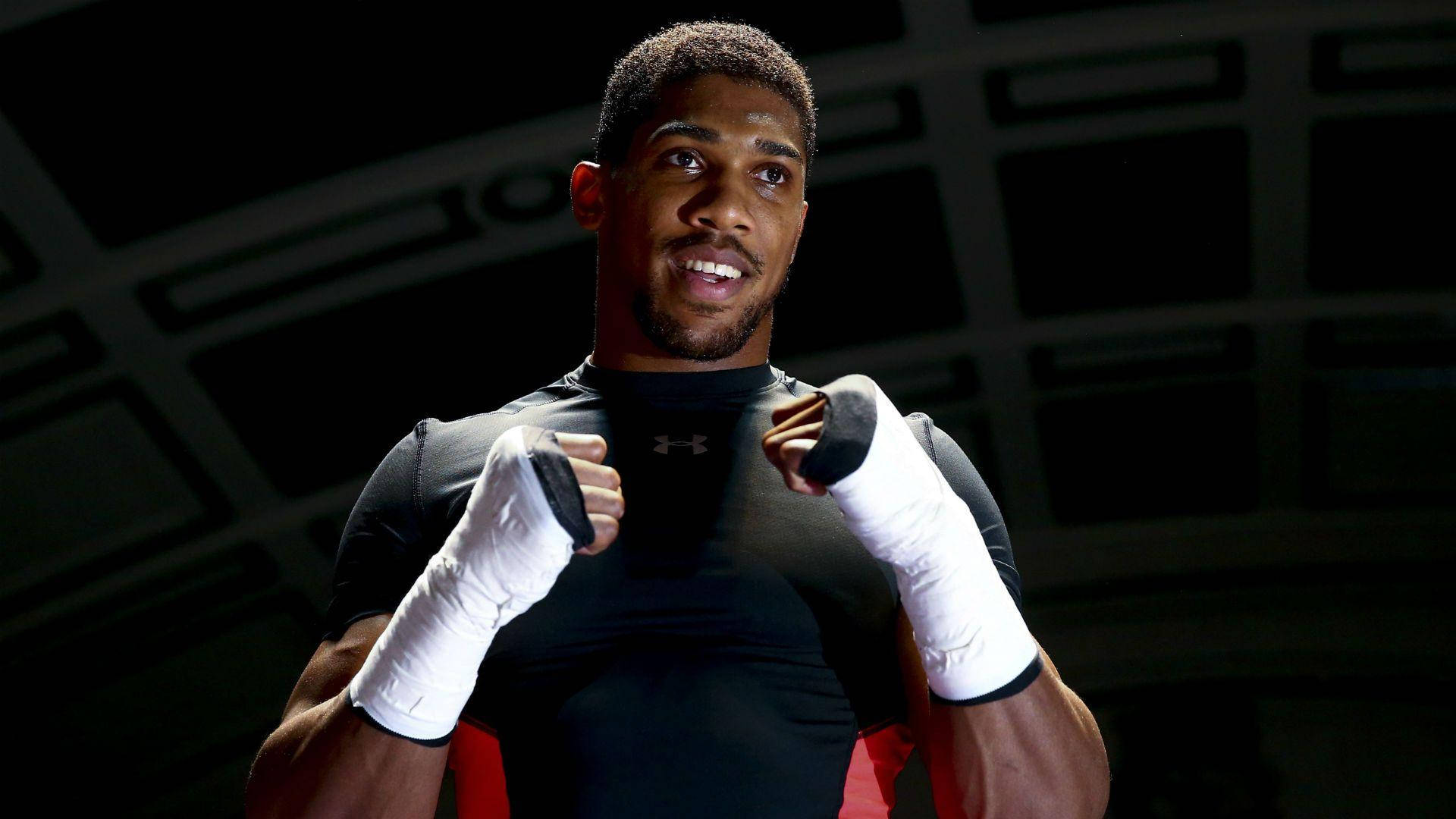 Anthony Joshua 桌布 壁紙 — 一位肌肉發達的拳擊手，戴著白色護手帶，身穿黑色襯衫，在深色幾何背景前自信地微笑，舉起拳頭。