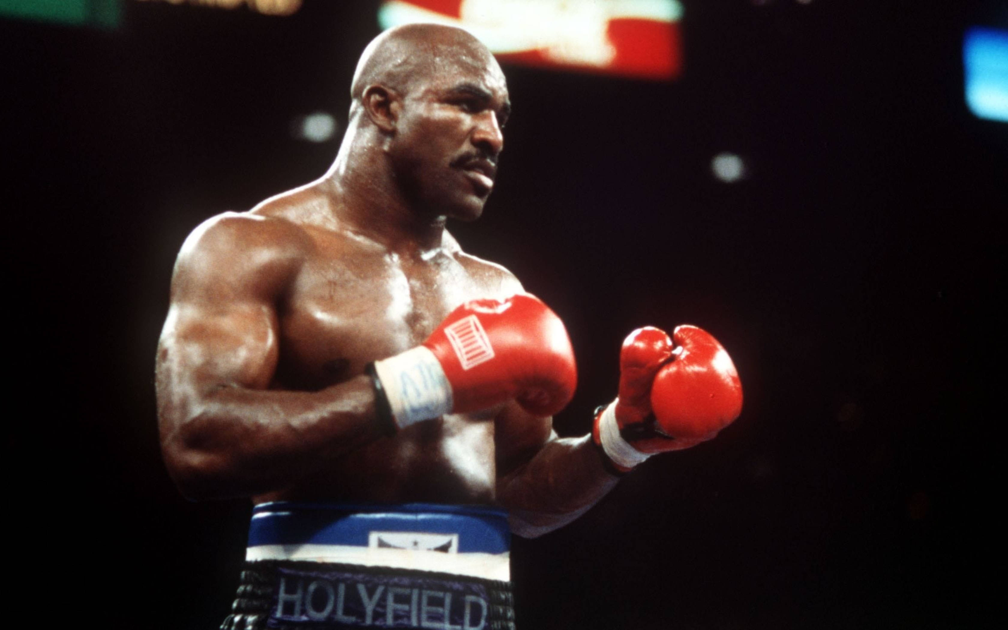 Boxing 桌布 壁紙 — 一位肌肉發達的拳擊手，戴著紅色手套和藍色短褲，在昏暗的擂台上專注地站立。