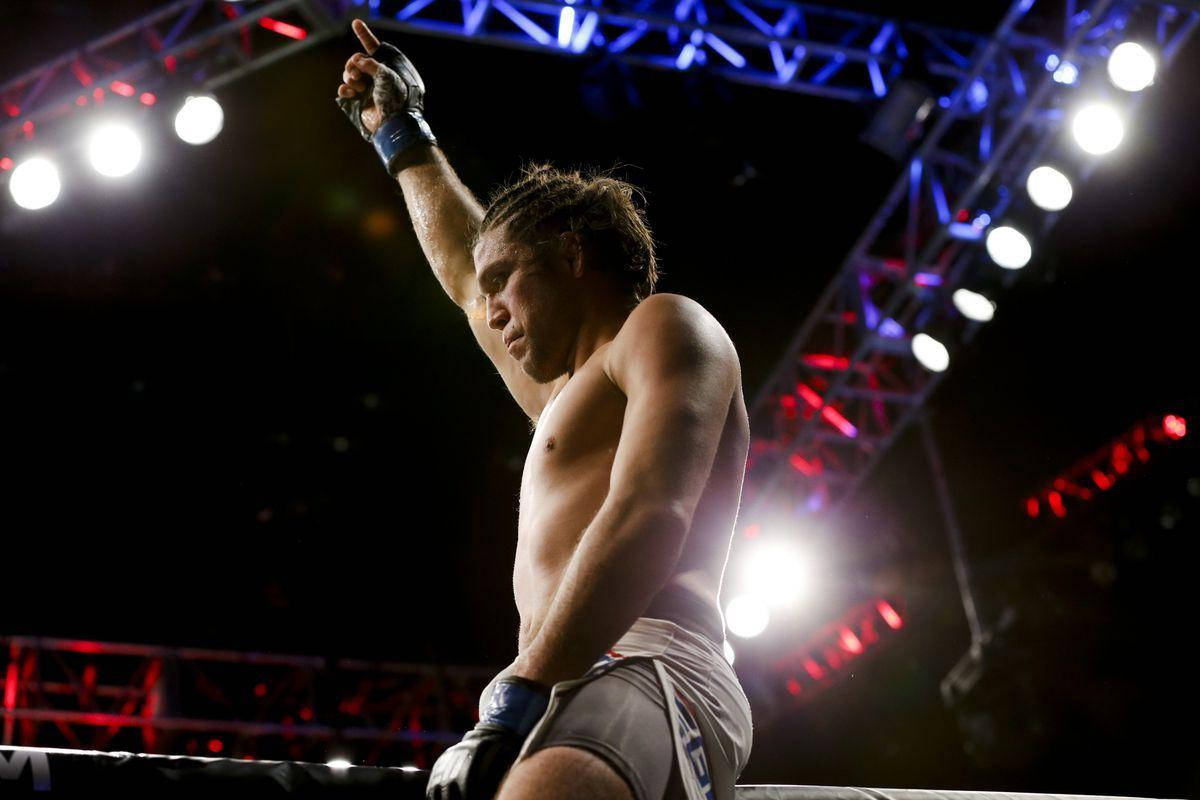 Brian Ortega 桌布 壁紙 — 一位肌肉發達的拳擊手在場館燈光下，於明亮照明的八角籠內舉起戴著手套的拳頭慶祝勝利。