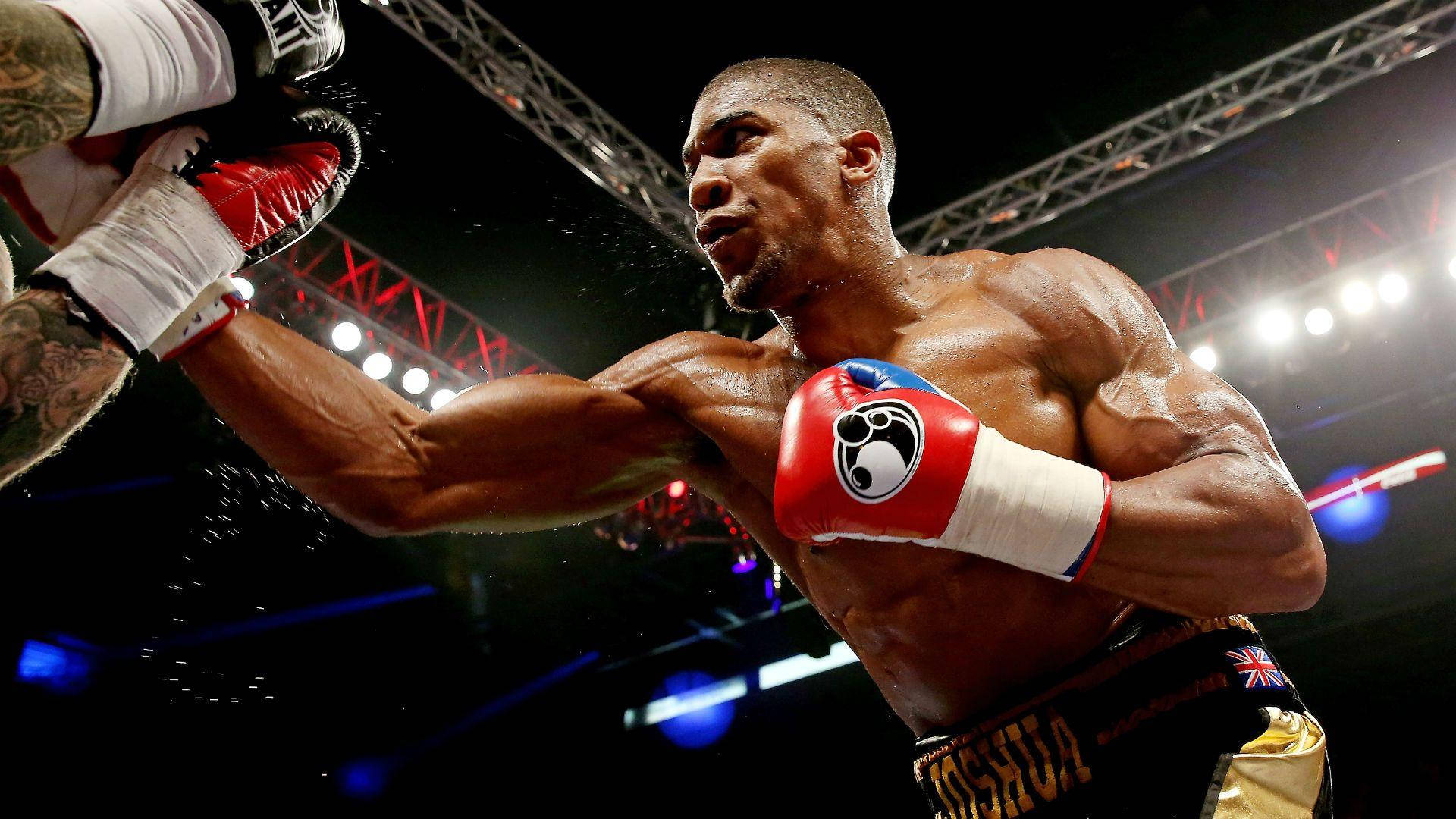 Anthony Joshua 桌布 壁紙 — 一位肌肉發達的拳擊手在燈光明亮的擂台上打出強力的右勾拳，汗水在空中飛濺。