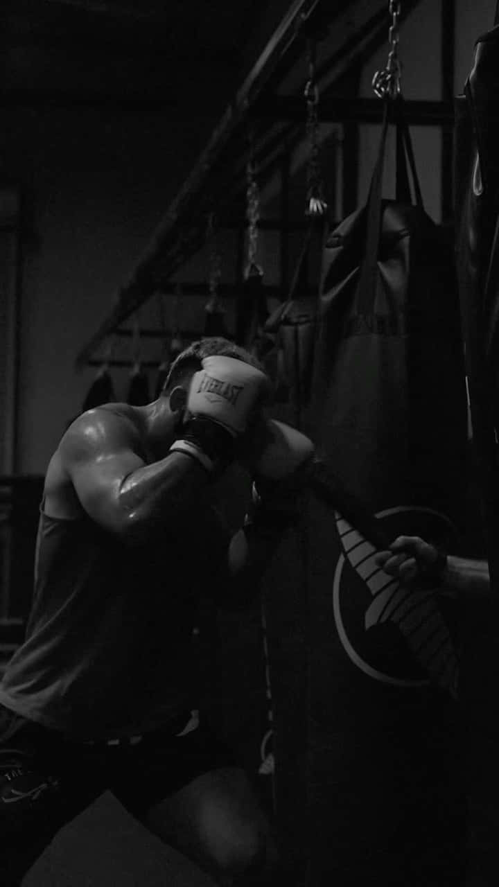 Boxing 桌布 壁紙 — 一位肌肉發達的拳擊手戴著手套，在燈光昏暗的健身房中專注地訓練，用力擊打沉重的沙袋。