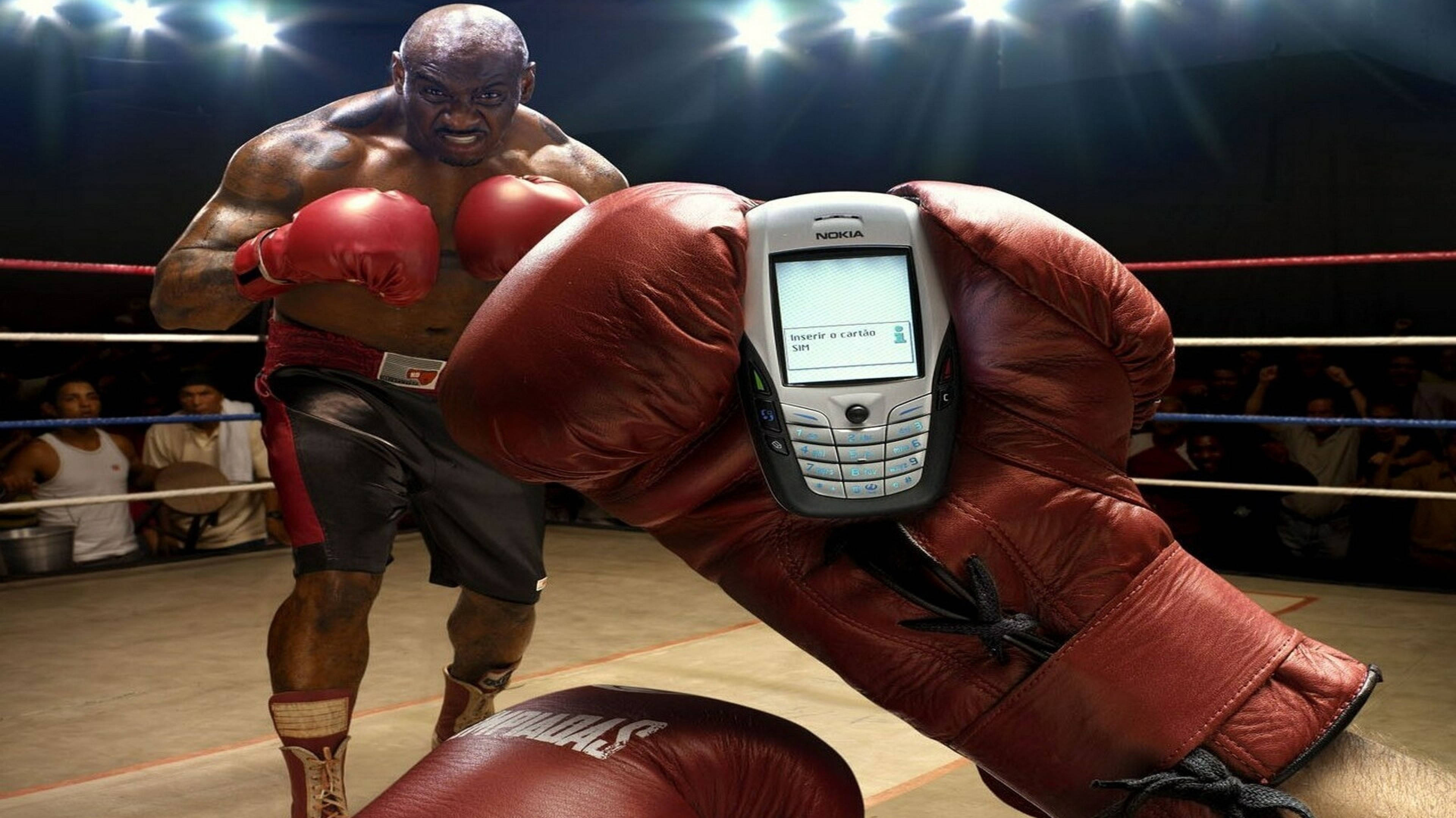 Cool Boxing 桌布 壁紙 — 一位肌肉發達的拳擊手戴著紅色手套，在燈光明亮的擂台上拳打諾基亞手機，觀眾在旁觀看。