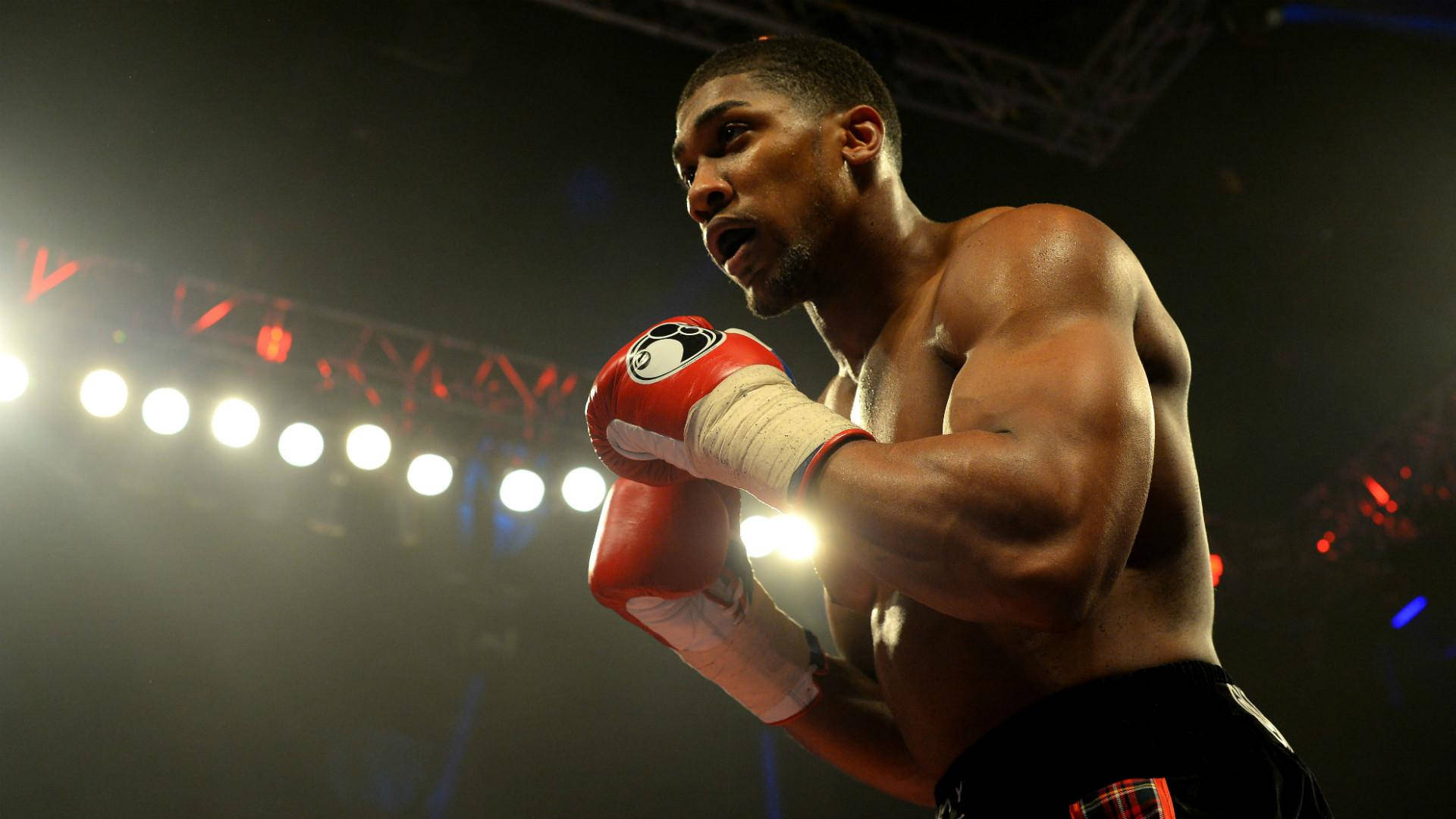 Anthony Joshua 桌布 壁紙 — 一位肌肉發達的拳擊手戴著紅色手套，在昏暗的擂台上，明亮的聚光燈下警覺地站立。