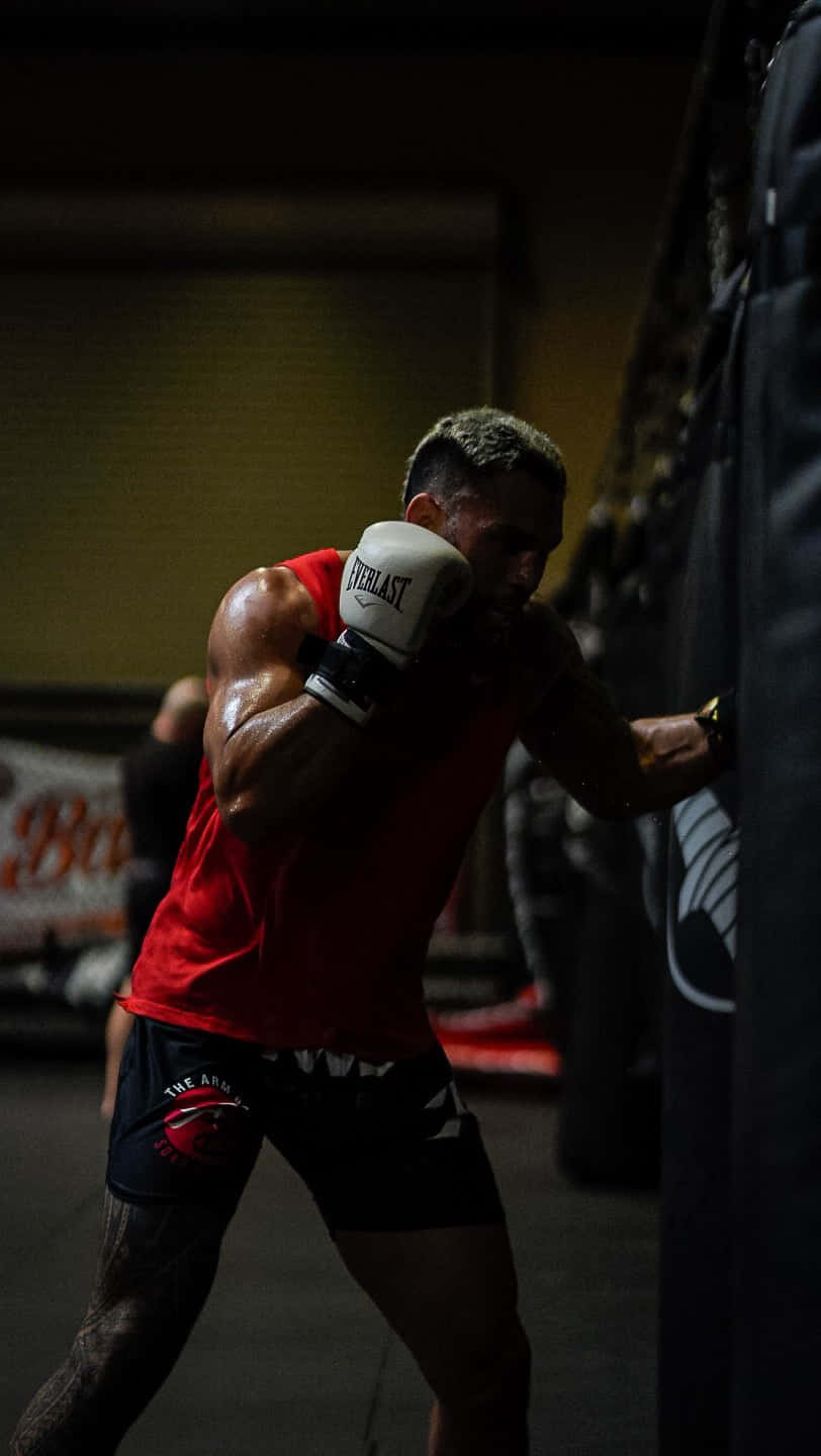 Boxing 桌布 壁紙 — 一位肌肉發達的拳擊手穿著紅色背心，在昏暗的健身房燈光下向沙包打出強力一拳。