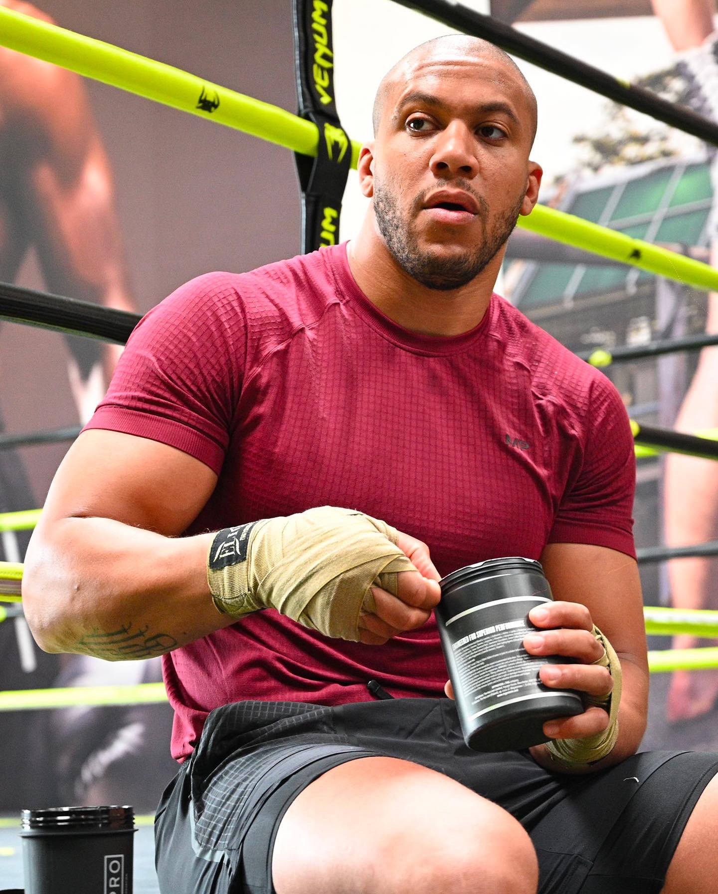 MMA 桌布 壁紙 — 一位肌肉發達的拳擊手穿著紅色襯衫坐在擂台上，專注地拿著一個黑色蛋白質容器。