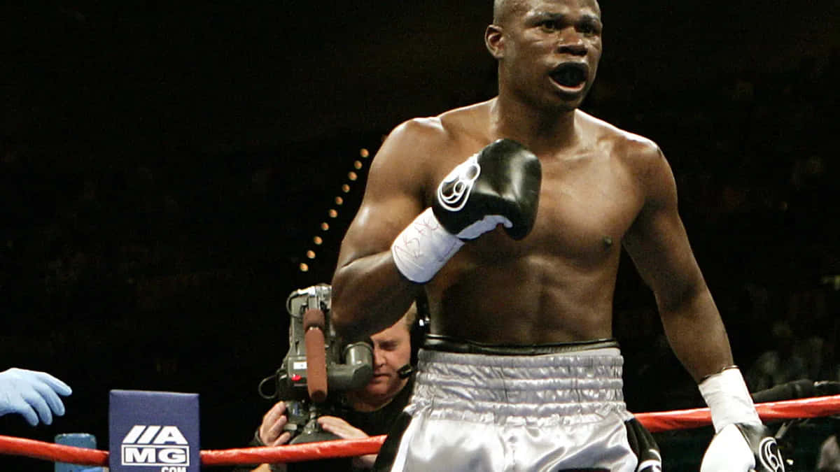 Boxing 桌布 壁紙 — 一位肌肉發達的拳擊手穿著銀色短褲，在燈光昏暗、有紅色繩索和攝影團隊在附近的擂台上勝利地咆哮。