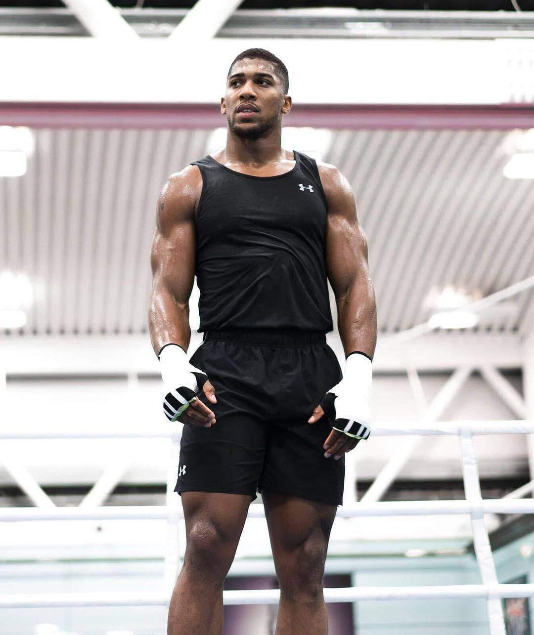 Anthony Joshua 桌布 壁紙 — 一位肌肉發達的拳擊手穿著黑色裝備和白色手套，專注地站在燈光明亮的擂台上。