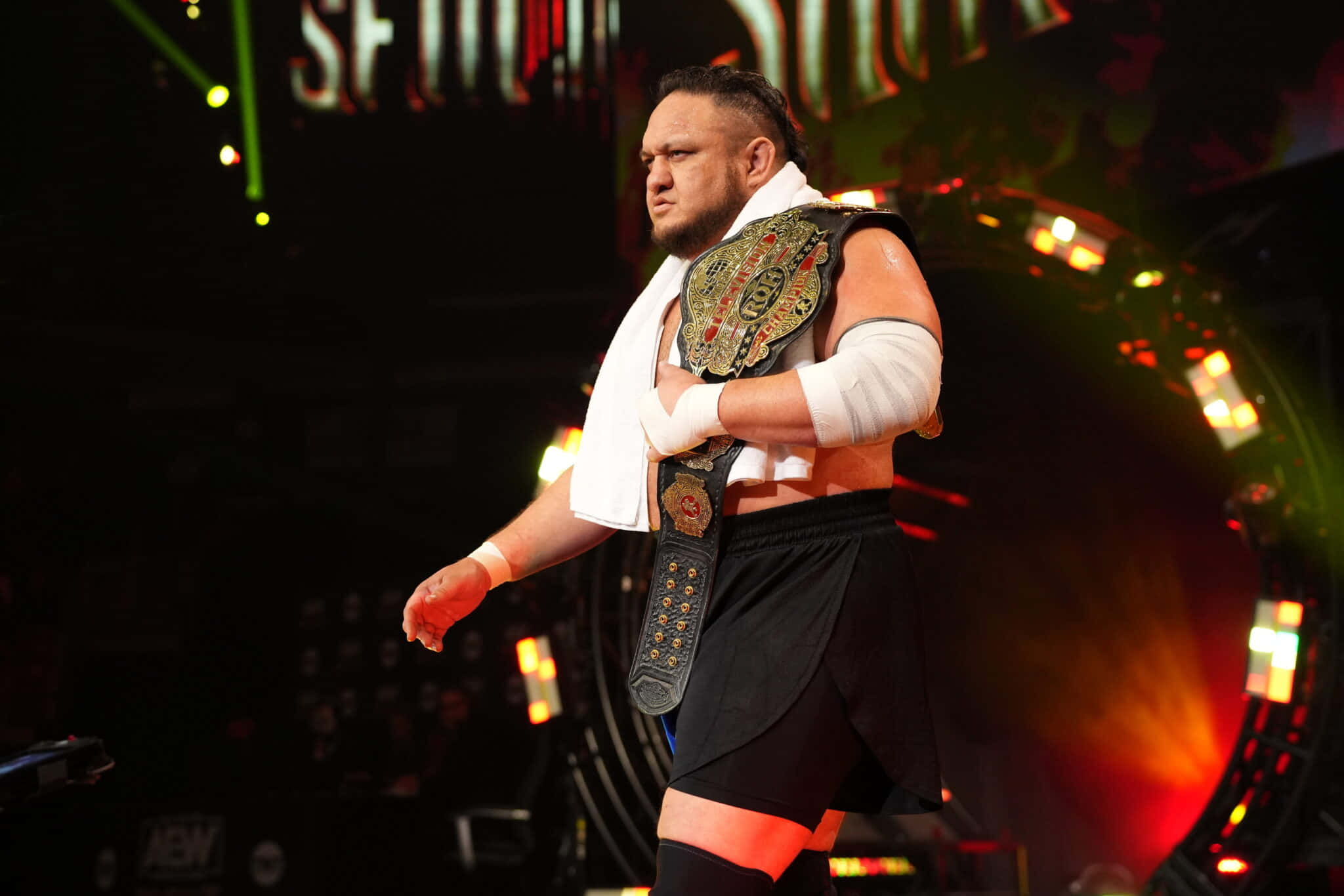 WWE 桌布 壁紙 — 一位肌肉發達的摔角手，冠軍腰帶搭在肩上，在明亮的舞台燈光下自信地大步前行。
