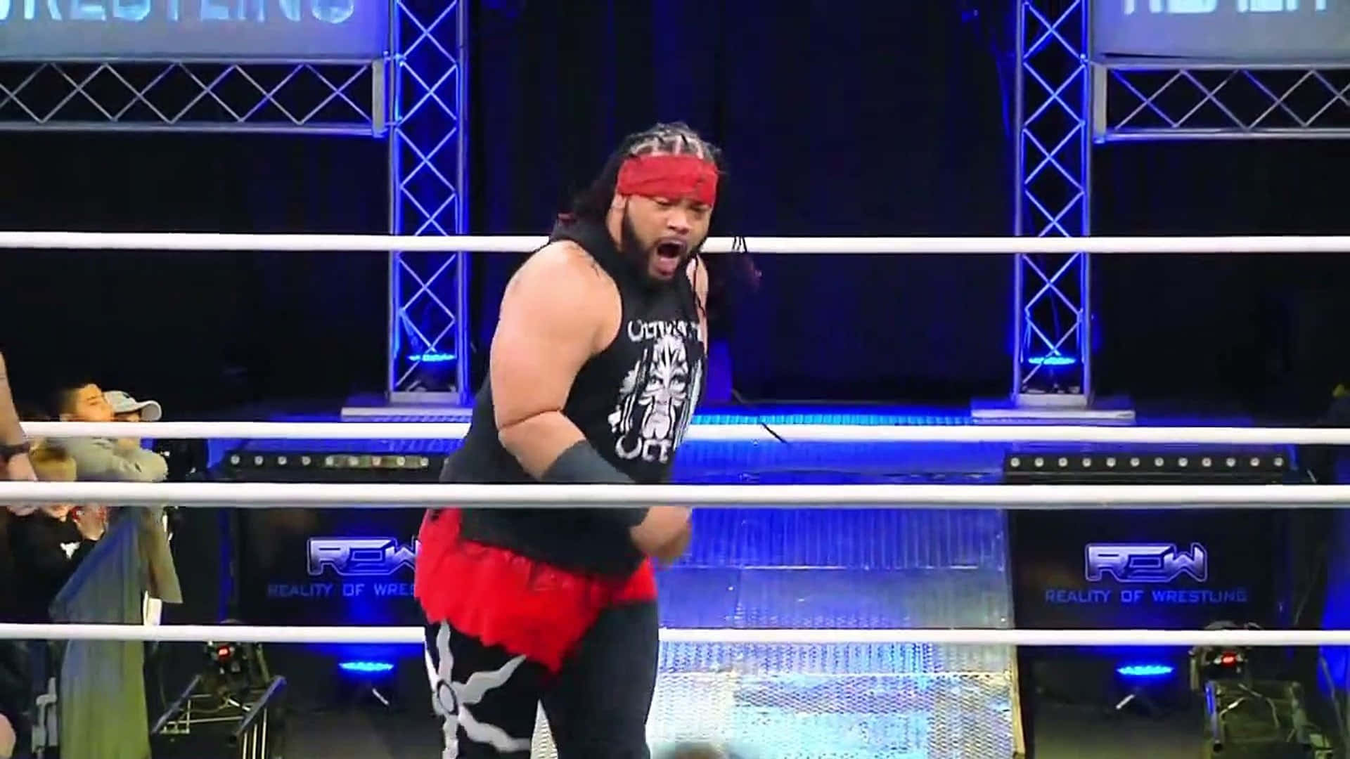WWE 桌布 壁紙 — 一位肌肉發達的摔角手，戴著紅色頭帶和黑色背心，在藍色舞台燈光照耀的擂台上咆哮。