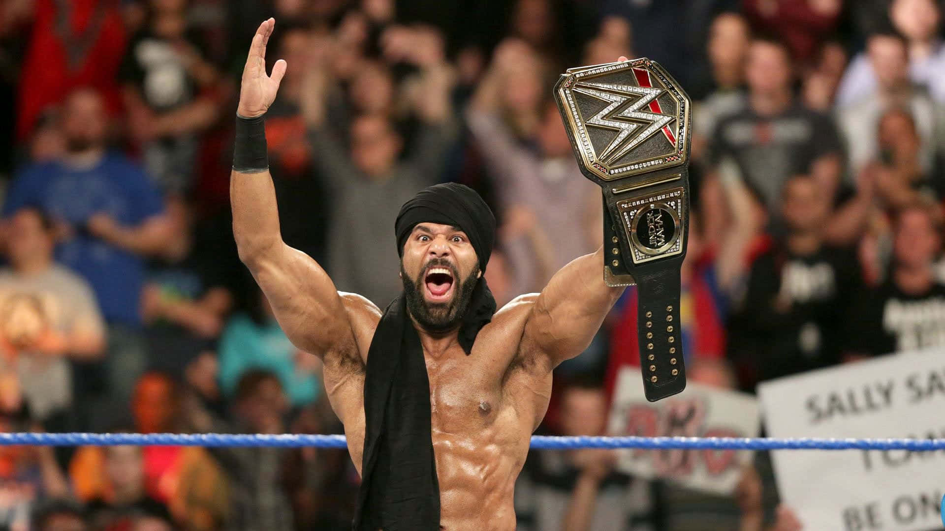 WWE 桌布 壁紙 — 一位肌肉發達的摔角手，戴著黑色頭巾和鬍鬚，在擠滿觀眾的場館中勝利地高舉WWE冠軍腰帶和拳頭。