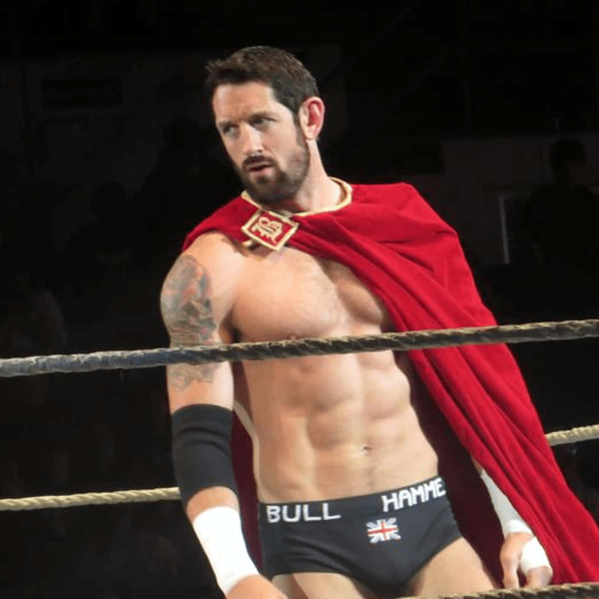 WWE 桌布 壁紙 — 一位肌肉發達的摔角手，披著紅色斗篷，手臂上有刺青，站在燈光明亮的摔角擂台上。