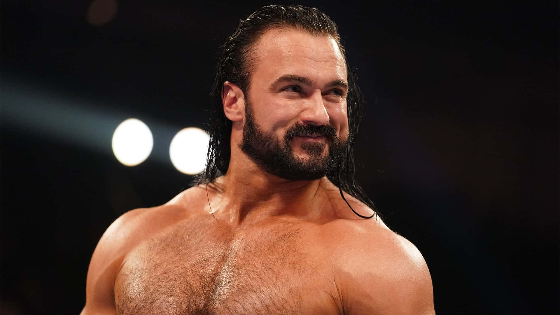WWE 桌布 壁紙 — 一位肌肉發達的摔角手，濕透的深色頭髮和鬍鬚，在明亮的競技場燈光下對著黑暗背景自信地微笑。