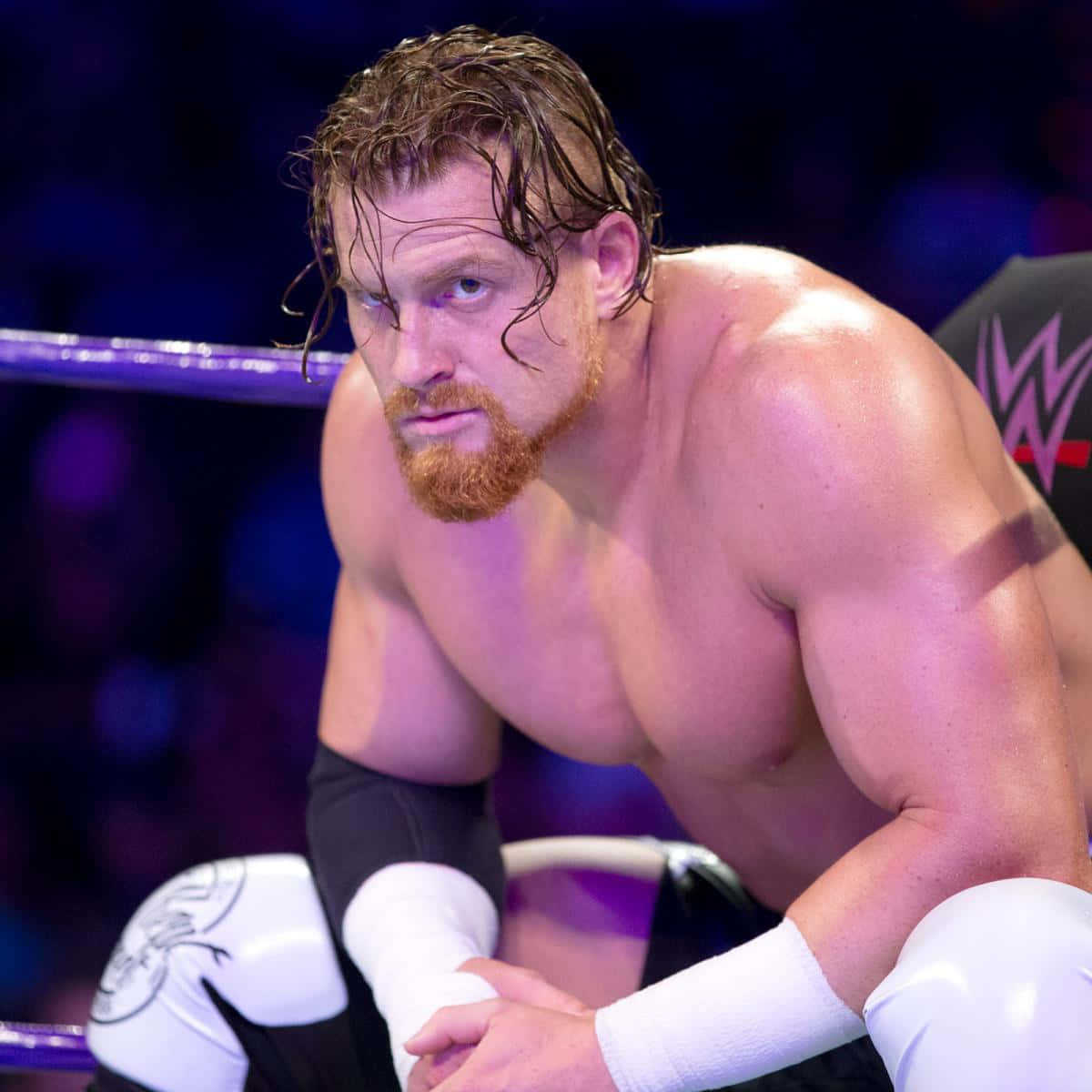 WWE 桌布 壁紙 — 一位肌肉發達的摔角手，濕髮紅鬚，在紫色燈光的擂台上蹲伏，神情專注。
