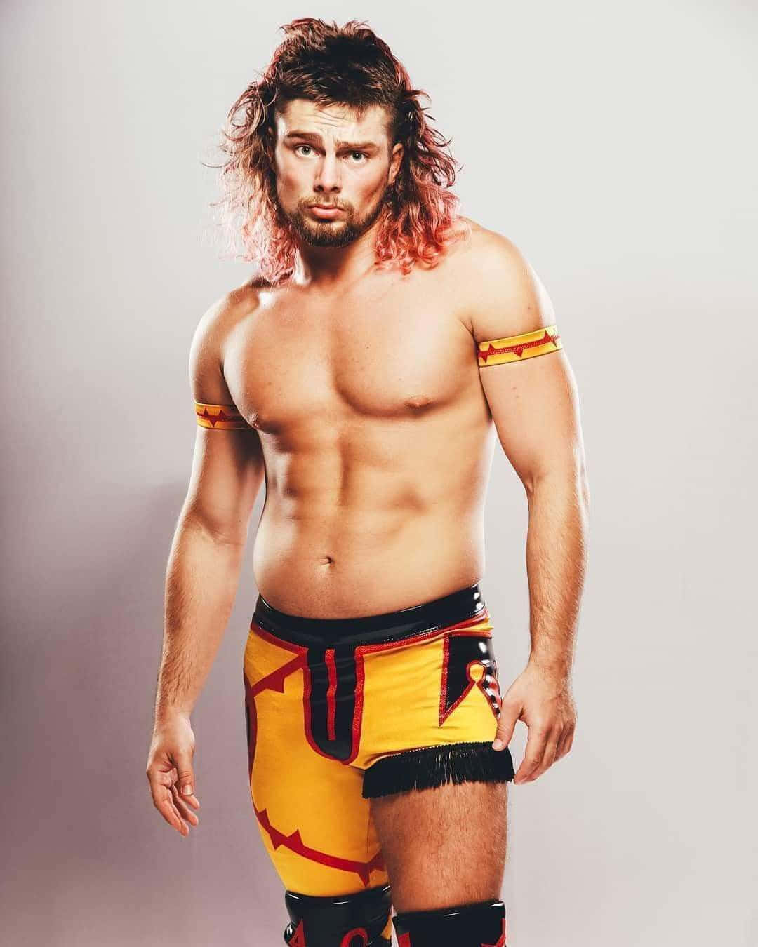 WWE 桌布 壁紙 — 一位肌肉發達的摔角手，留著長長的紅髮，穿著黃色短褲，在純灰色工作室背景前自信地擺姿勢。
