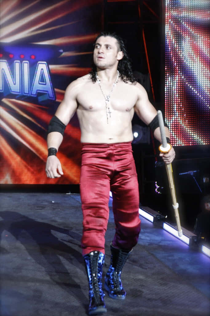 WWE 桌布 壁紙 — 一位肌肉發達的摔角手，身穿紅色緞面褲子和藍色靴子，手持權杖自信地走過燈光明亮的舞台。