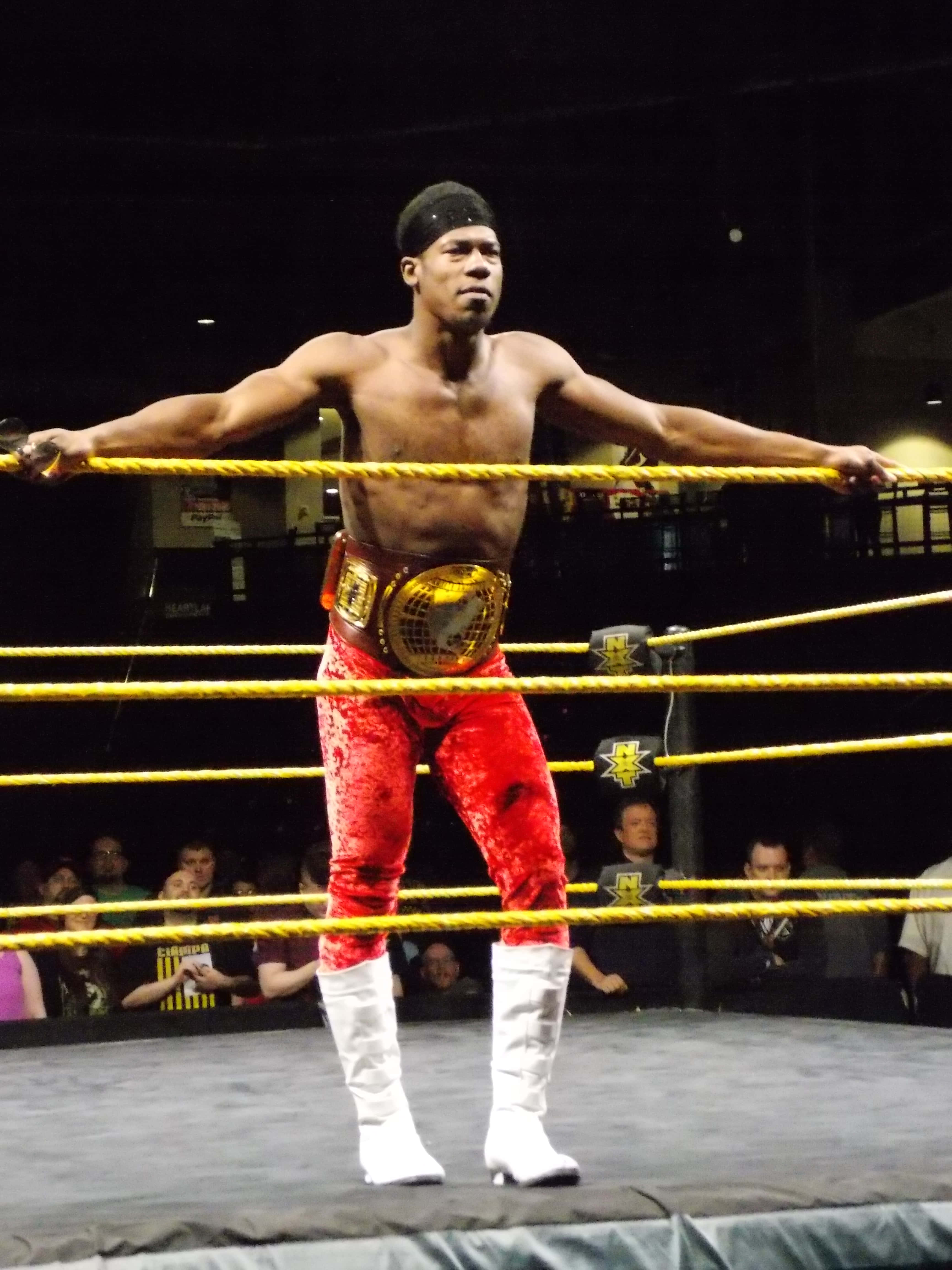 WWE 桌布 壁紙 — 一位肌肉發達的摔角手，身穿紅色褲子和白色靴子，自信地站在摔角擂台上，手握繩索，腰間繫著冠軍腰帶。