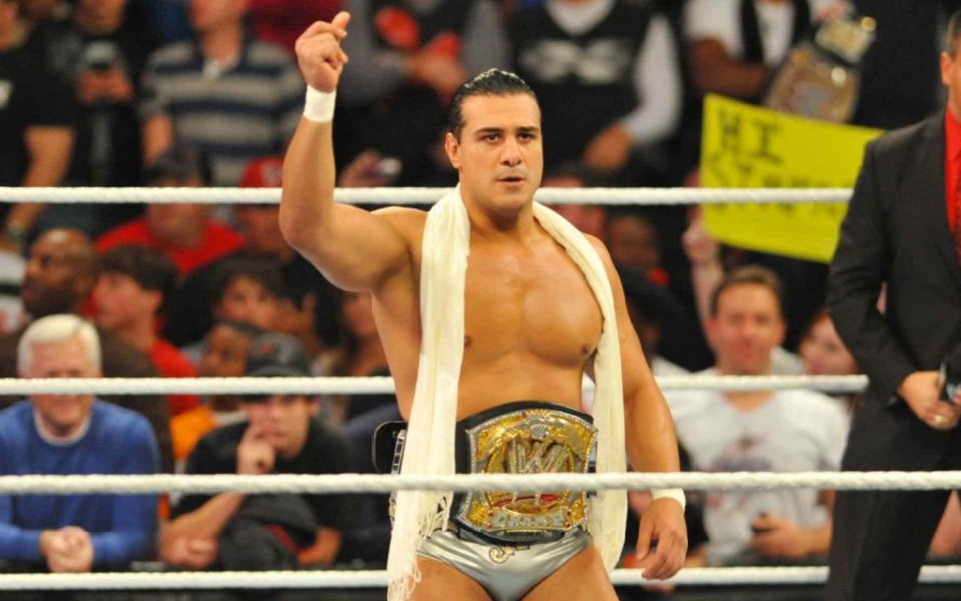 WWE 桌布 壁紙 — 一位肌肉發達的摔角手，身穿銀色短褲和冠軍腰帶，在摔角擂台內高舉拳頭，慶祝勝利，四周是歡呼的粉絲。