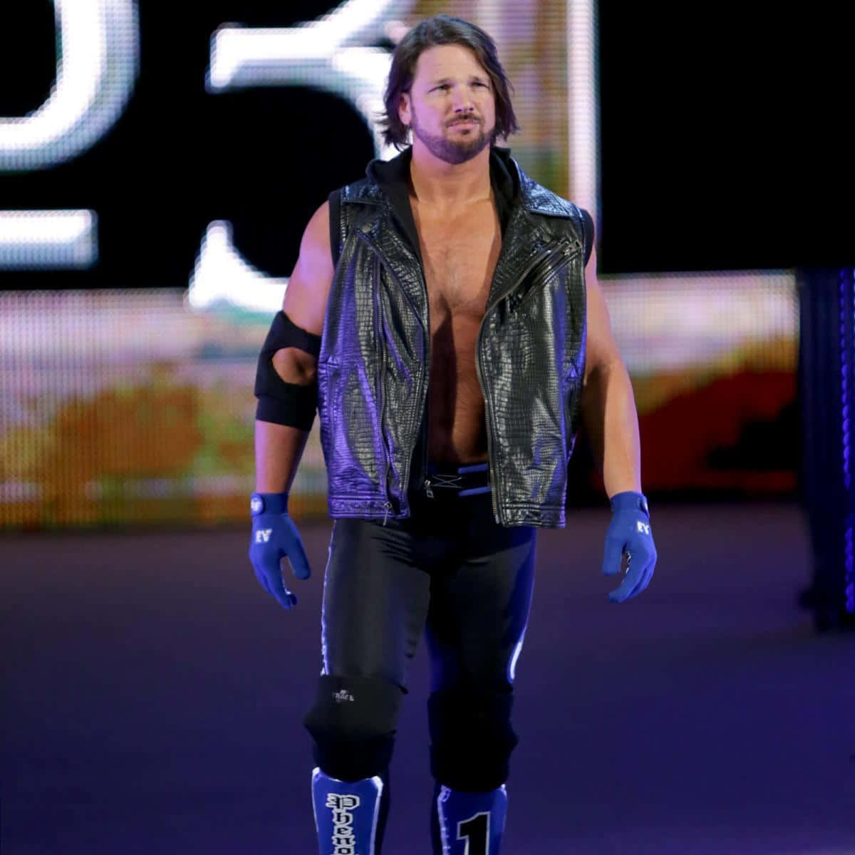 WWE 桌布 壁紙 — 一位肌肉發達的摔角手，身穿黑色皮背心和藍色手套，自信地走過燈光昏暗的競技場舞台。