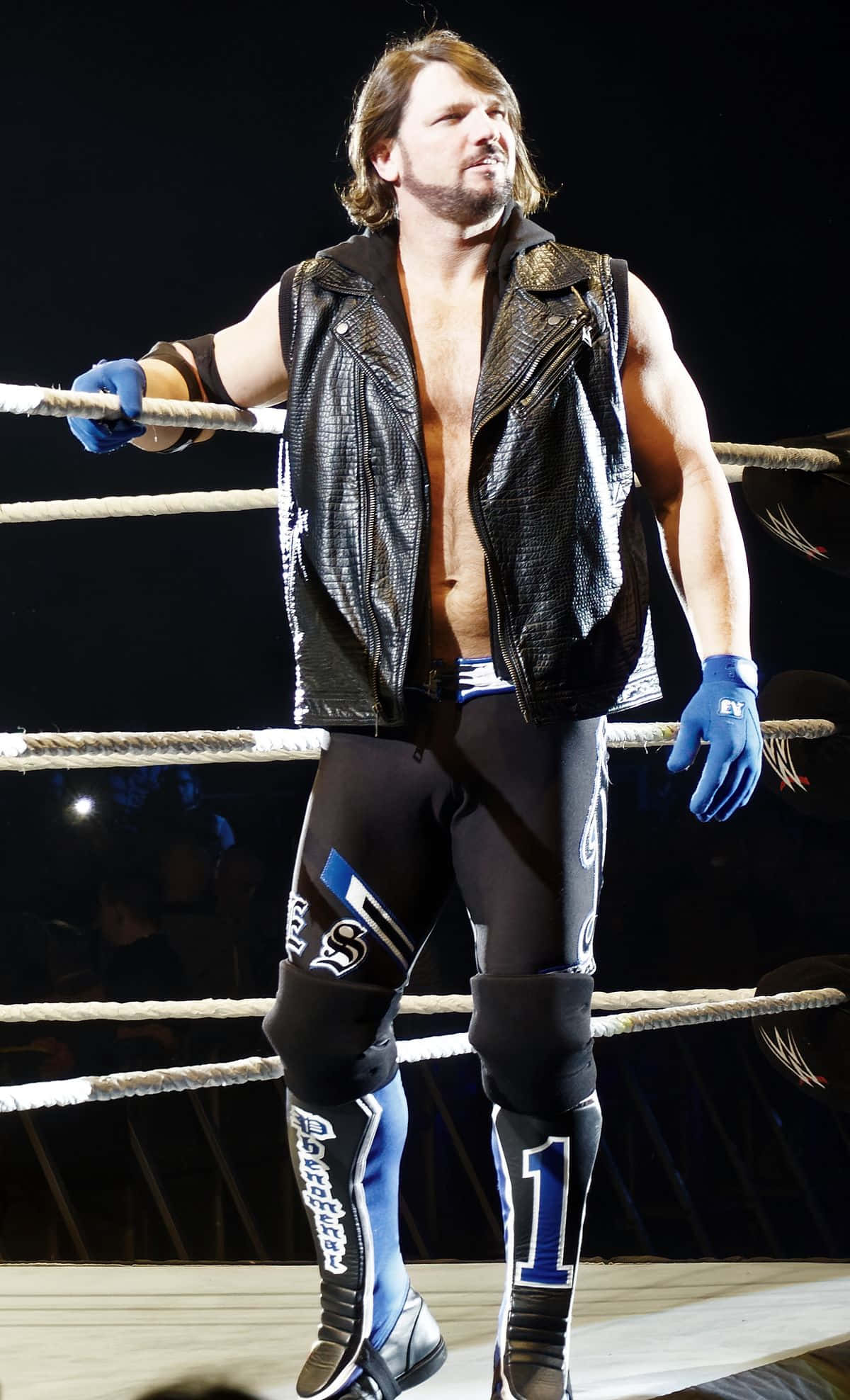 WWE 桌布 壁紙 — 一位肌肉發達的摔角手，身穿黑色皮背心和藍色手套，在明亮的場館燈光下自信地站在摔角擂台內。