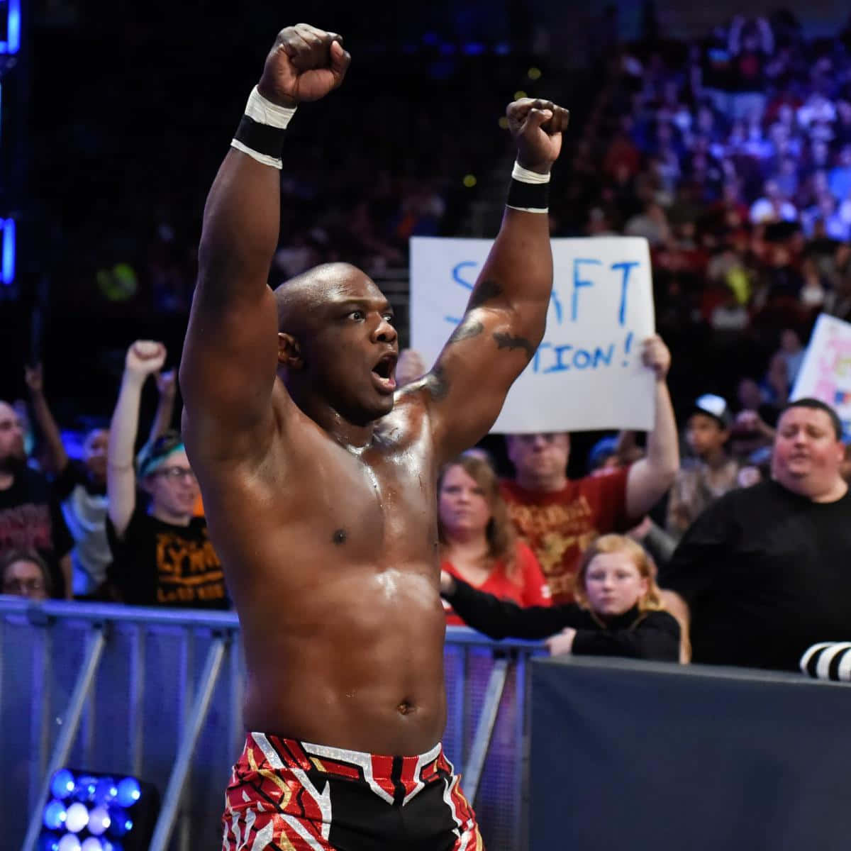 WWE 桌布 壁紙 — 一位肌肉發達的摔角手在擂台上勝利地高舉雙拳，身後歡呼的人群舉著標語牌。