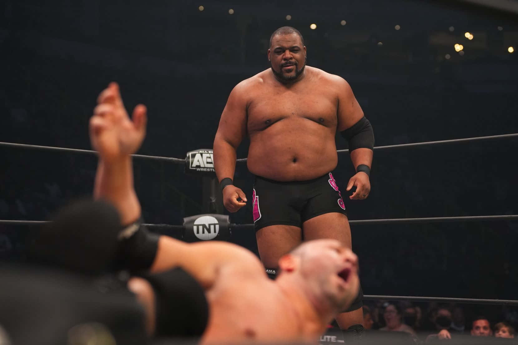 Aew 桌布 壁紙 — 一位肌肉發達的摔角手在擂台上勝利地站著，而他的對手則被擊敗，雙臂高舉以示認輸。