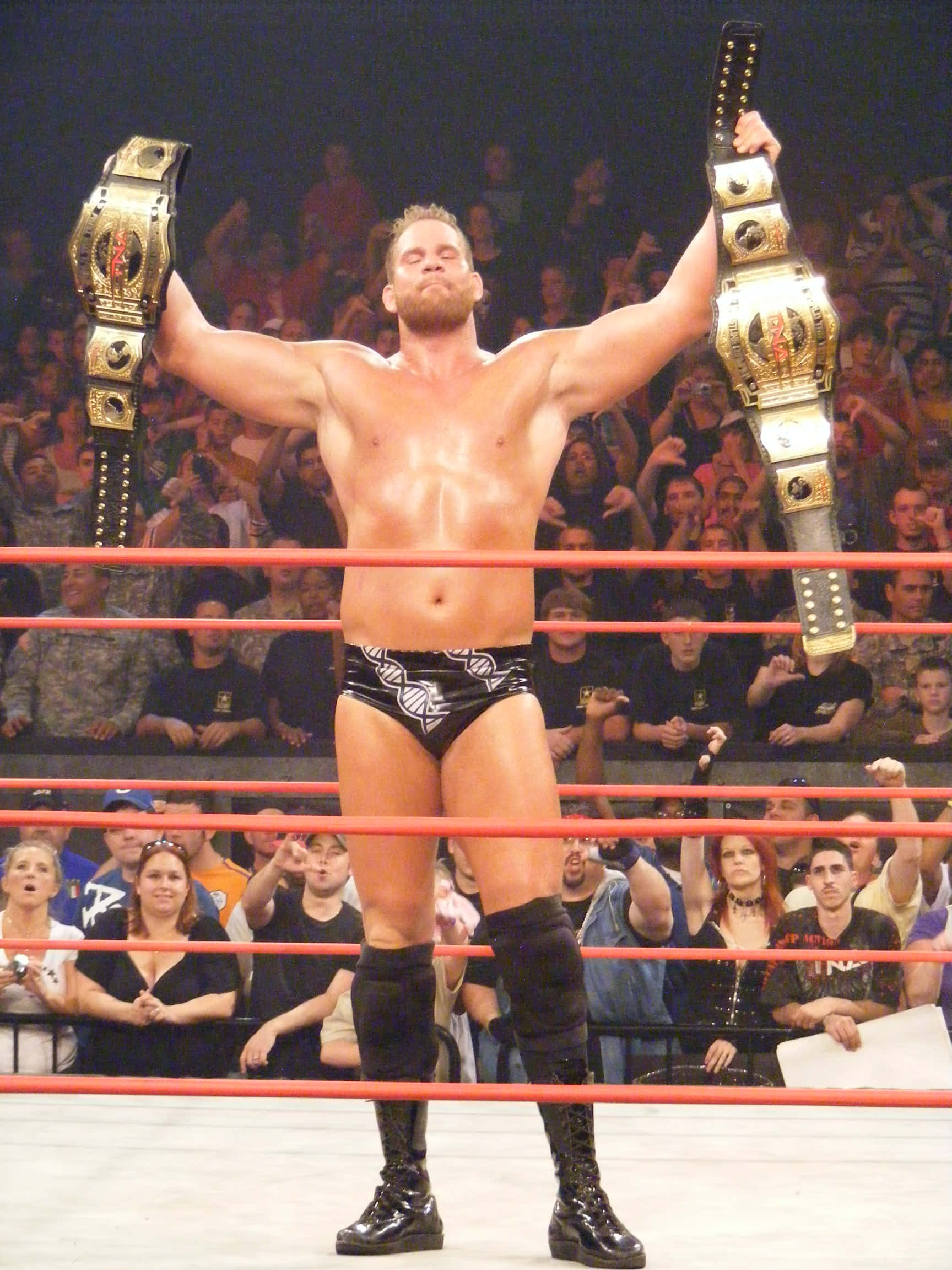 WWE 桌布 壁紙 — 一位肌肉發達的摔角手在擂台上高舉兩條冠軍腰帶，身後興奮的人群為他歡呼。