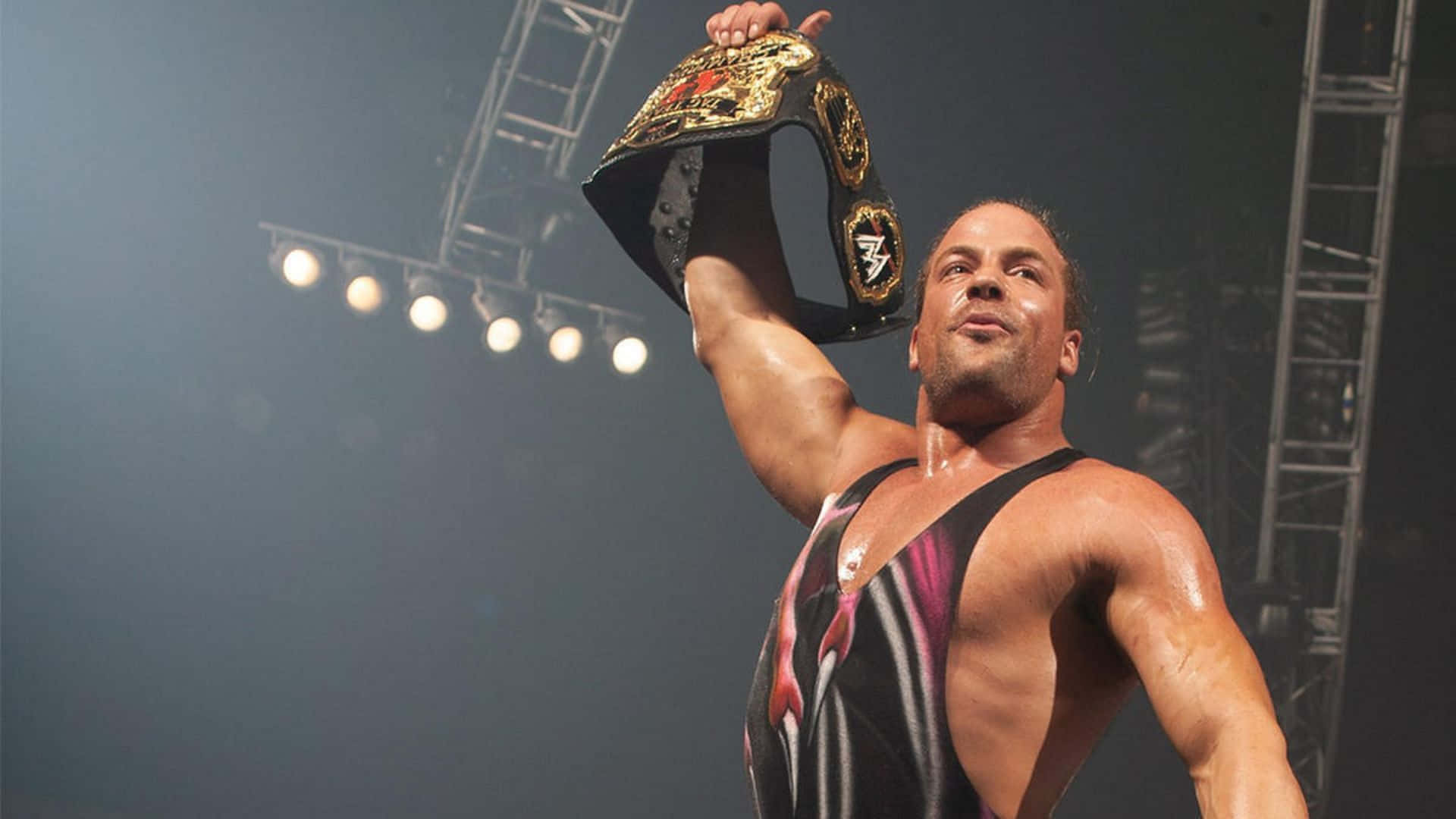 WWE 桌布 壁紙 — 一位肌肉發達的摔角手在明亮的場館燈光下勝利地高舉冠軍腰帶。