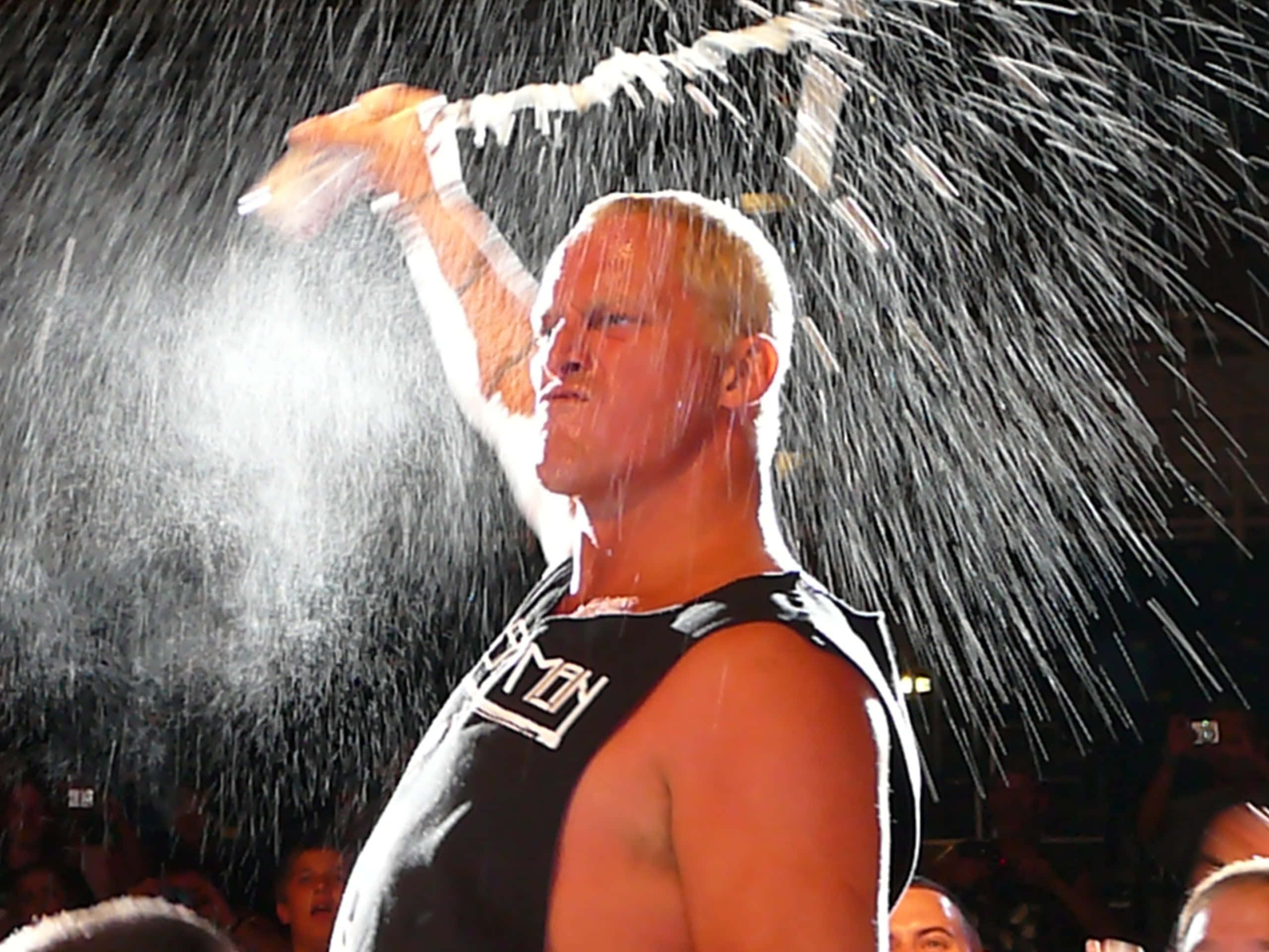 WWE 桌布 壁紙 — 一位肌肉發達的摔角手在明亮的場館燈光下將水澆在頭上，下方是歡呼的人群。