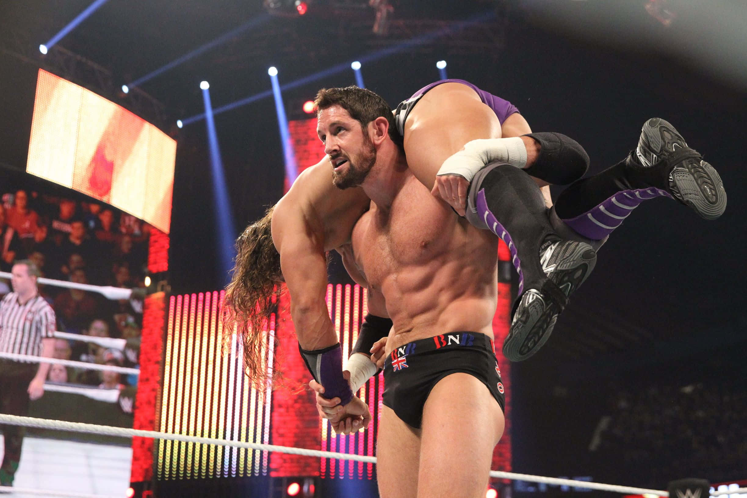 WWE 桌布 壁紙 — 一位肌肉發達的摔角手在明亮的場館燈光下，於摔角台上將對手高舉過頭。