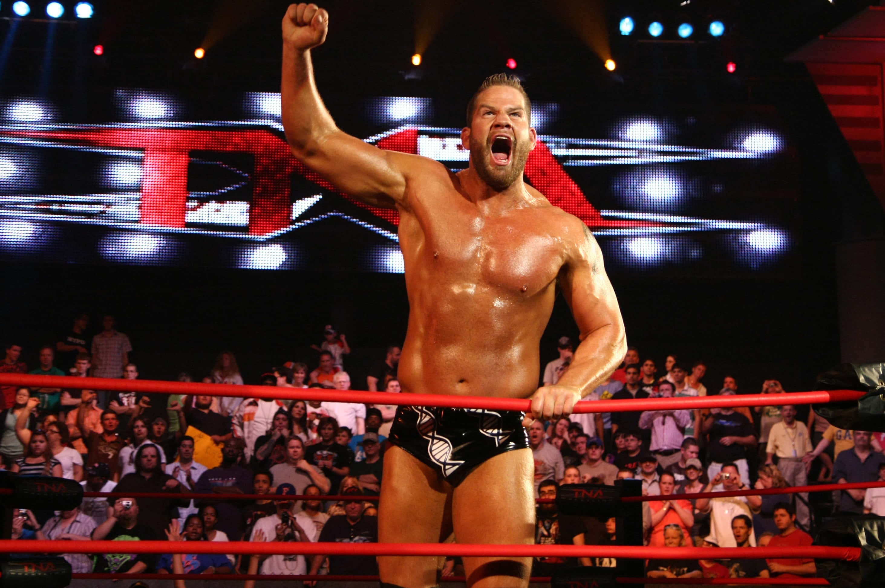 WWE 桌布 壁紙 — 一位肌肉發達的摔角手在明亮的場館燈光下，於紅色繩索的擂台上發出勝利的吼叫。