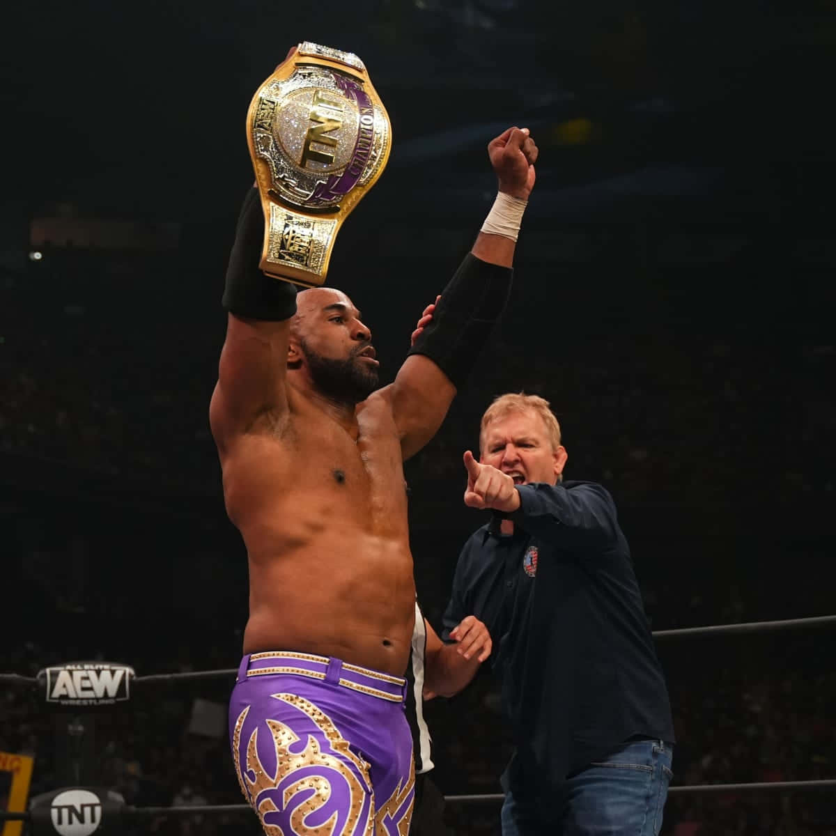 Wrestling 桌布 壁紙 — 一位肌肉發達的摔角手在燈光明亮的擂台上，勝利地將一條金色和紫色的冠軍腰帶高舉過頭，旁邊一位穿藍色襯衫的男子在歡呼。