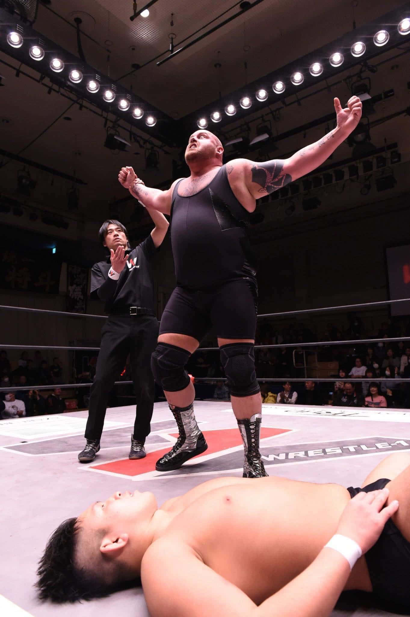 WWE 桌布 壁紙 — 一位肌肉發達的摔角手在燈光明亮的摔角場內，勝利地高舉雙臂，慶祝擊敗倒下的對手。