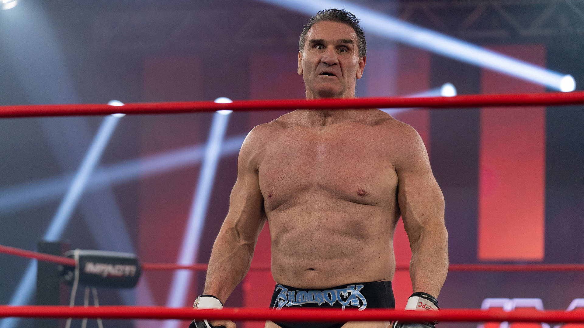 WWE 桌布 壁紙 — 一位肌肉發達的摔角手在燈光明亮的擂台上，紅繩和聚光燈在頭頂，神情專注。