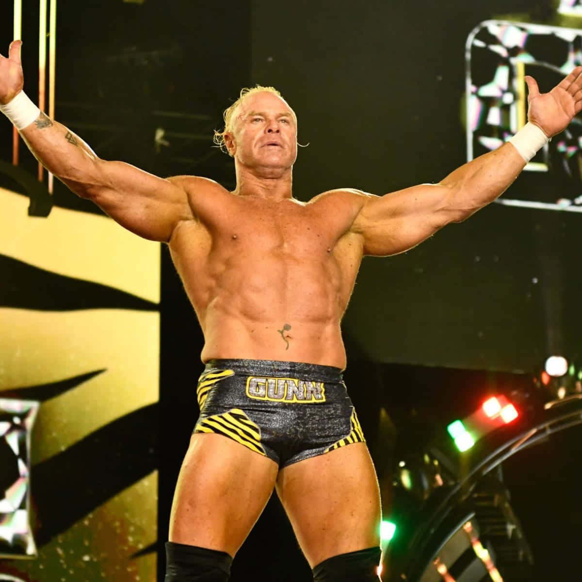 WWE 桌布 壁紙 — 一位肌肉發達的摔角手在燈光明亮的舞台上高舉雙臂，穿著黑色和黃色的短褲，腰帶上印有「GUNN」字樣。