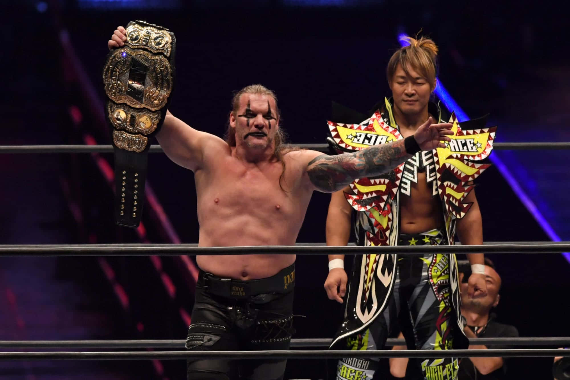Hiroshi Tanahashi 桌布 壁紙 — 一位肌肉發達的摔角手在燈光明亮的擂台上高舉冠軍腰帶，慶祝勝利，另一位摔角手站在他旁邊。