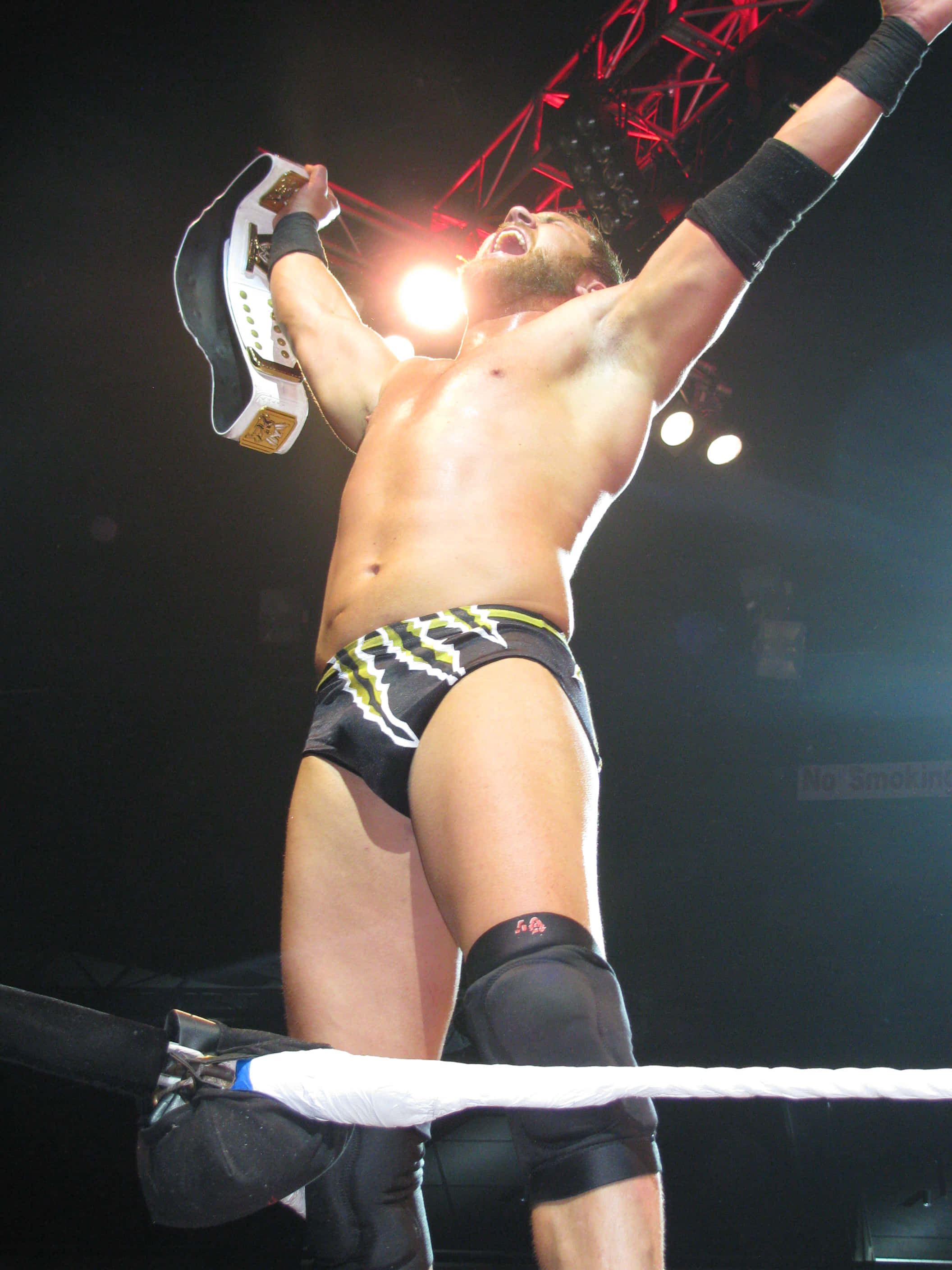 WWE 桌布 壁紙 — 一位肌肉發達的摔角手在燈光明亮的擂台上，勝利地將冠軍腰帶高舉過頭。