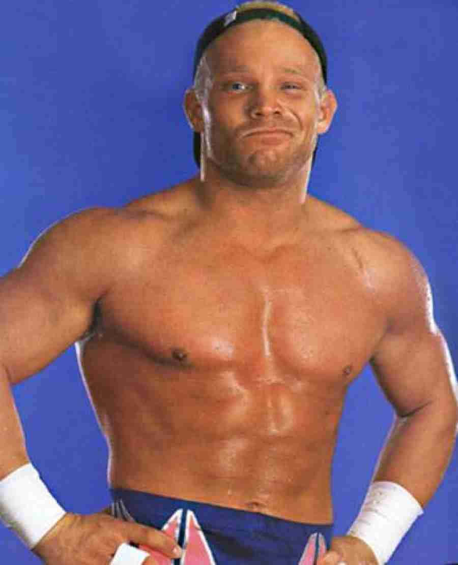 Crash Holly 桌布 壁紙 — 一位肌肉發達的摔角手帶著自信的笑容，赤裸上身站在純藍色背景前，穿著藍色短褲和白色腕帶。