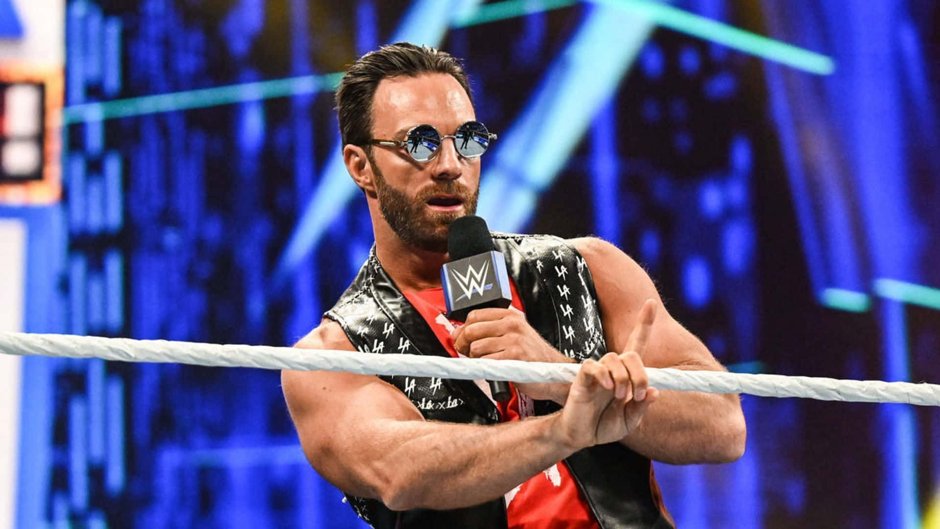 WWE 桌布 壁紙 — 一位肌肉發達的摔角手戴著太陽眼鏡和黑色背心，抓著擂台繩索，手持WWE麥克風，背景是藍色燈光的競技場。