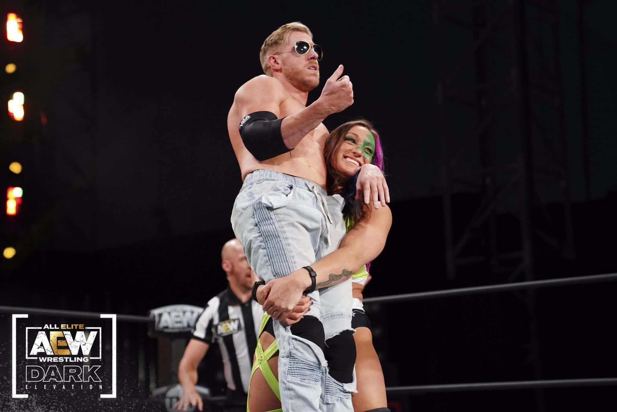 Aew 桌布 壁紙 — 一位肌肉發達的摔角手戴著太陽眼鏡豎起大拇指，同時被一位微笑的女摔角手穿著霓虹裝備擁抱在摔角場內。