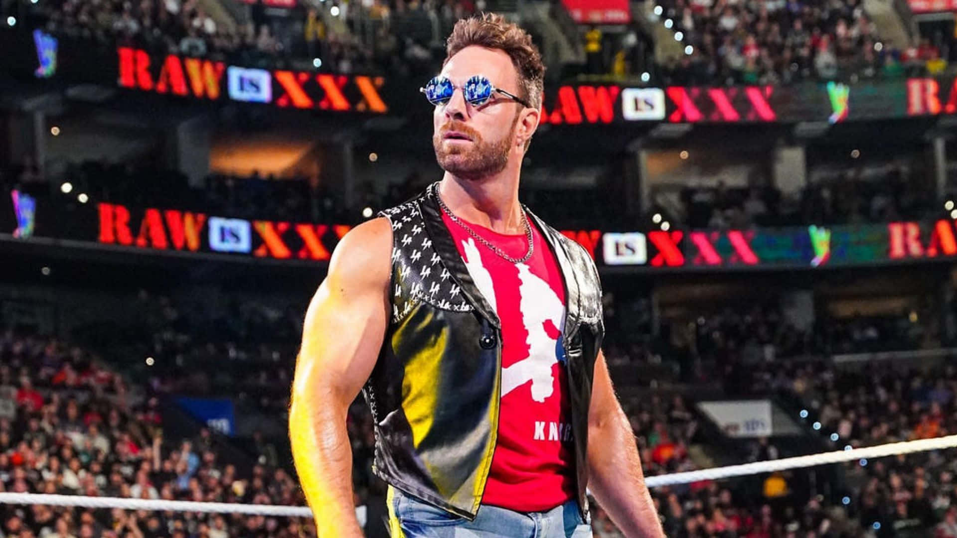 WWE 桌布 壁紙 — 一位肌肉發達的摔角手戴著太陽眼鏡和皮背心，自信地站在摔角擂台上，身後是座無虛席的場館。