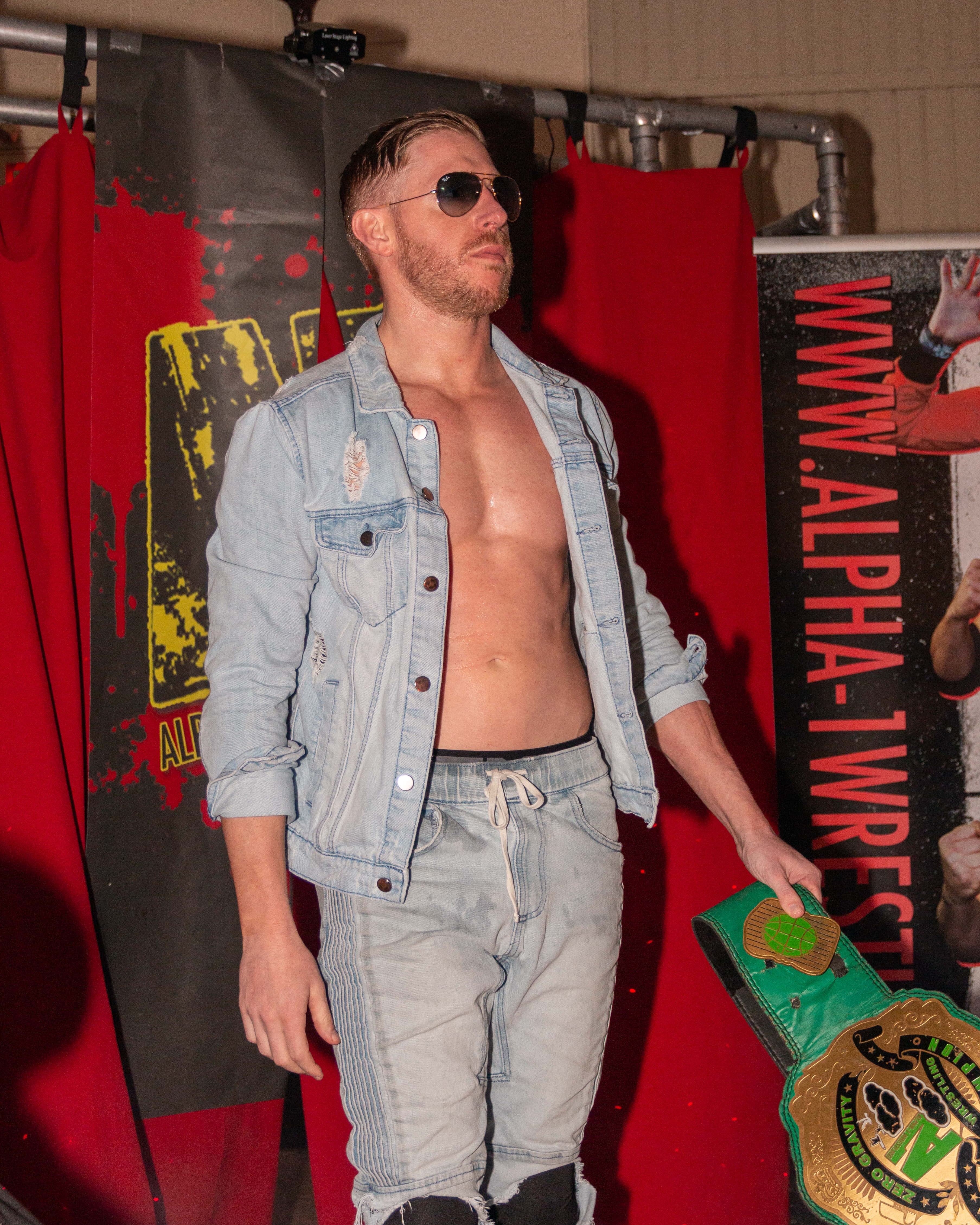 Aew 桌布 壁紙 — 一位肌肉發達的摔角手戴著太陽眼鏡，身穿開胸牛仔外套，在紅黑背景下拿著綠色冠軍腰帶。