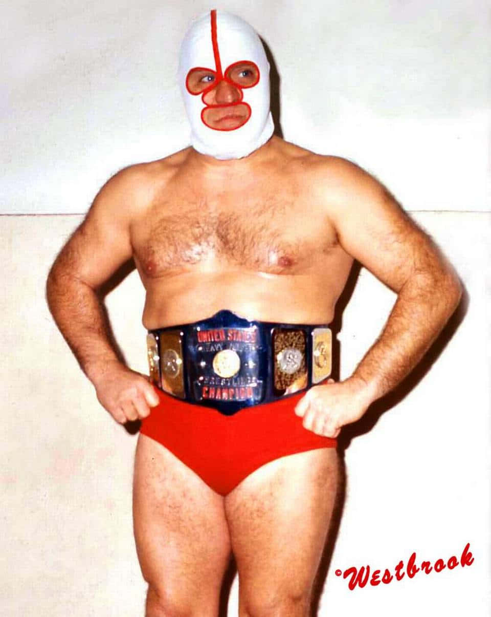 WWE 桌布 壁紙 — 一位肌肉發達的摔角手戴著帶有紅色點綴的白色面具，自信地站在素牆前，腰間繫著冠軍腰帶，穿著紅色短褲。