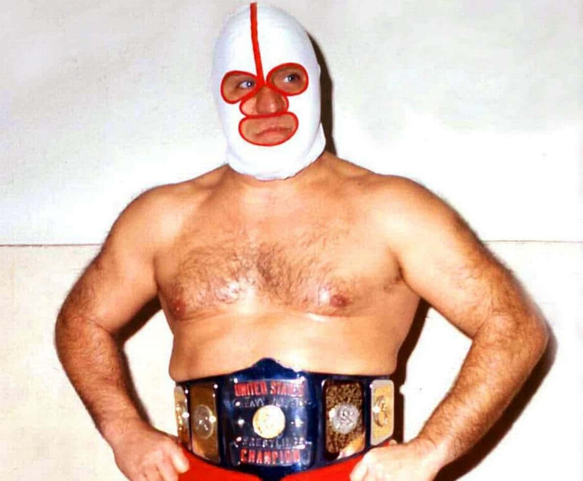 WWE 桌布 壁紙 — 一位肌肉發達的摔角手戴著白色和紅色的面具，雙手叉腰自信地站著，腰間繫著冠軍腰帶。