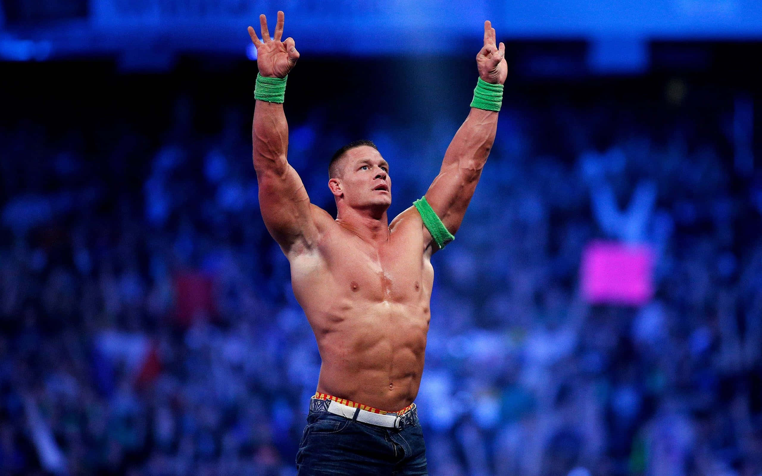 WWE 桌布 壁紙 — 一位肌肉發達的摔角手戴著綠色護腕，在藍色場館燈光下，於模糊的歡呼人群前高舉雙臂，展現勝利姿態。