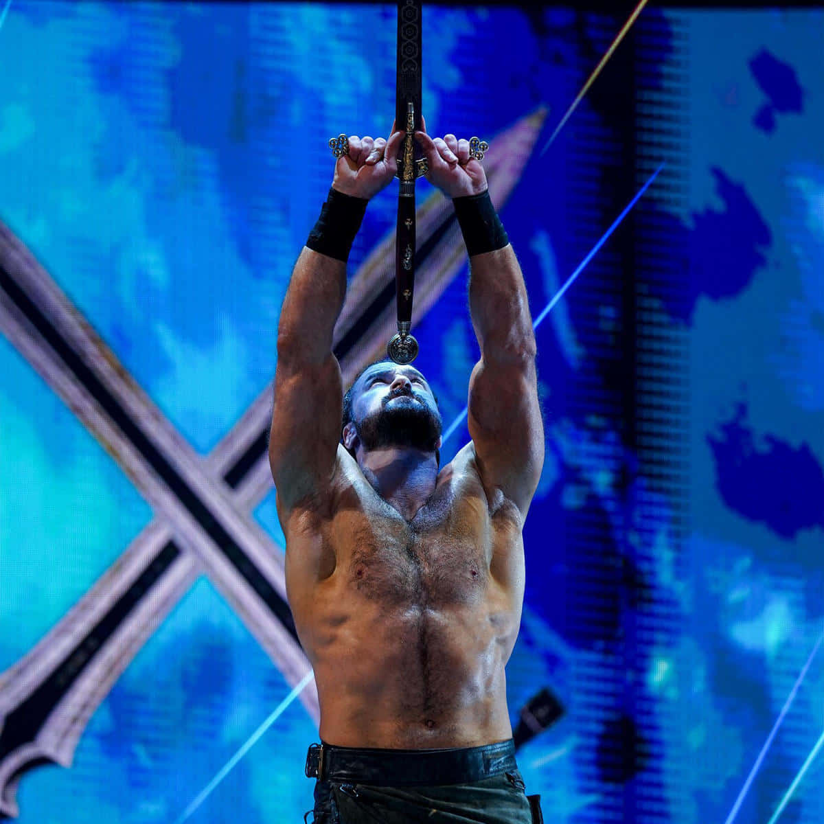 WWE 桌布 壁紙 — 一位肌肉發達的摔角手掛在劍上，懸浮於帶有X設計的藍色燈光舞台上方。