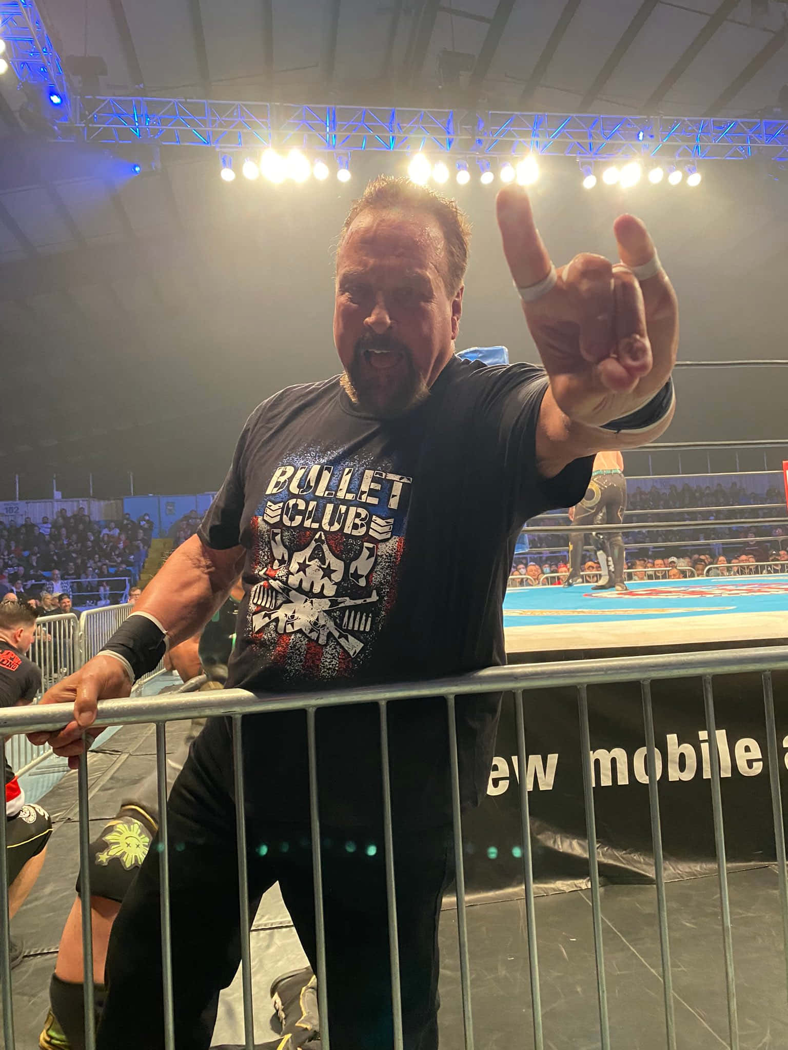 摔角 桌布 壁紙 — 一位肌肉發達的摔角手穿著 Bullet Club 襯衫，在燈光明亮的競技場金屬護欄後面向粉絲做出搖滾手勢。